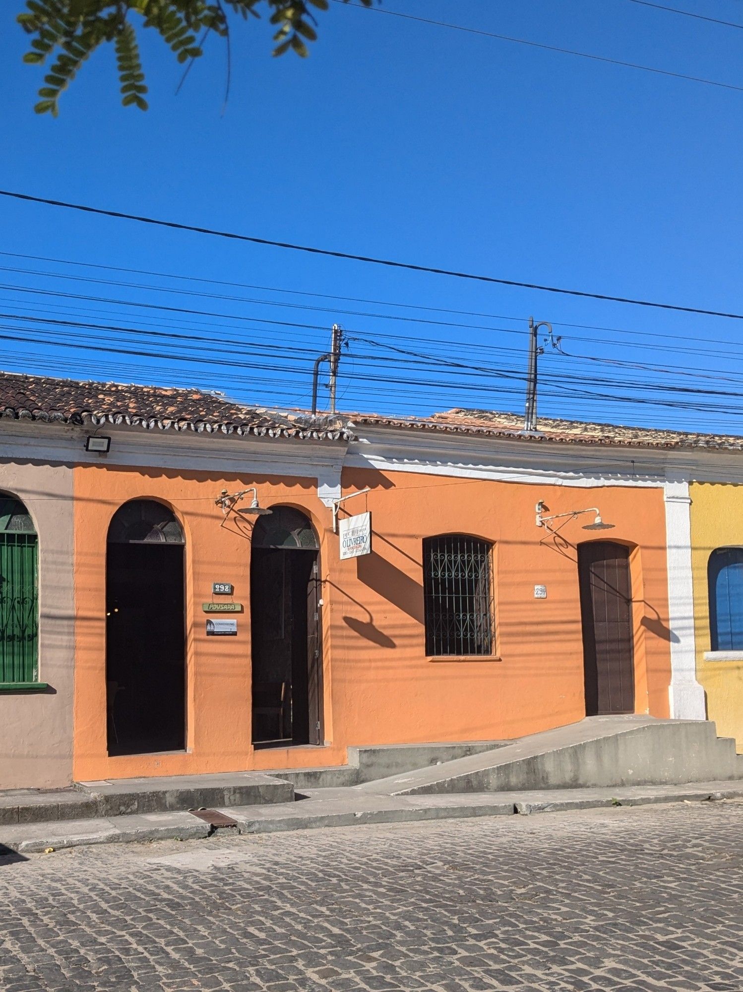 fachada do sebo "o livreiro", localizado na rua do mangue, em porto seguro, bahia. a foto foi tirada do outro lado da rua, então na parte de baixo é possível ver os paralelepípedos por onde passam os carros e pedestres. na parte de cima, podemos ver o céu bem azul e os fios de eletricidade altos que abastecem as casas, além de umas folhinhas de árvore no canto esquerdo da foto. a fachada do sebo em foco é laranja e tem o estilo arquitetônico clássico da cidade, as casinhas com pé direito alto, portas e janelas de madeira e uns poucos detalhes em branco. é possível ver uns pedaços das casas vizinhas também, amarela com detalhes azuis do lado direito e cinza com detalhes verdes do lado esquerdo