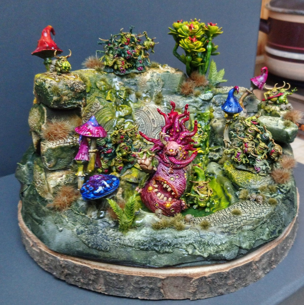 Front view of a painted small diorama. It's a ruin garden ith a small pond and lots of mushrooms and plants. A Beast of Nurgle is sitting in the pond and Nurglings are tending to it.