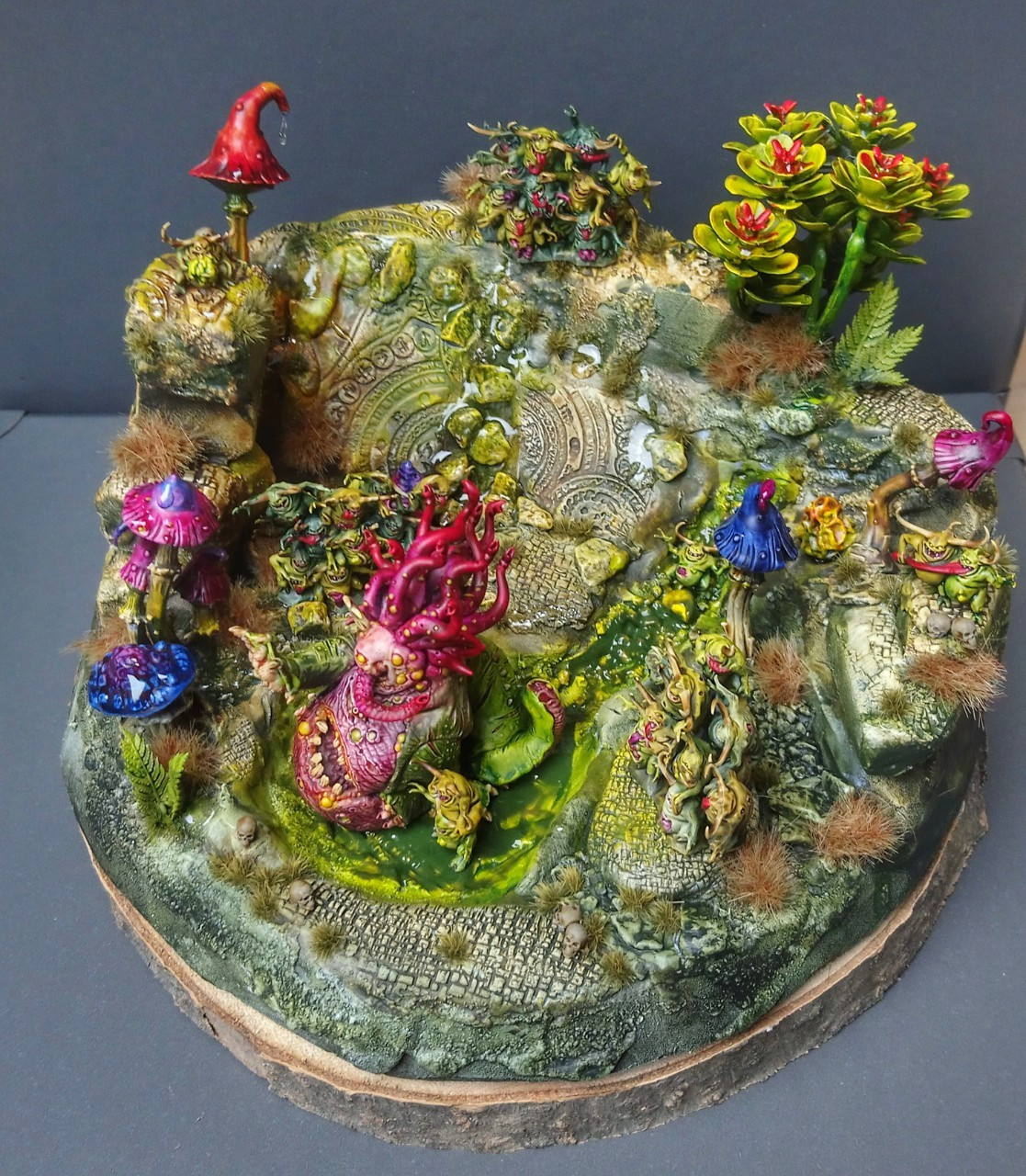 Top view of a painted small diorama. It's a ruin garden ith a small pond and lots of mushrooms and plants. A Beast of Nurgle is sitting in the pond and Nurglings are tending to it.