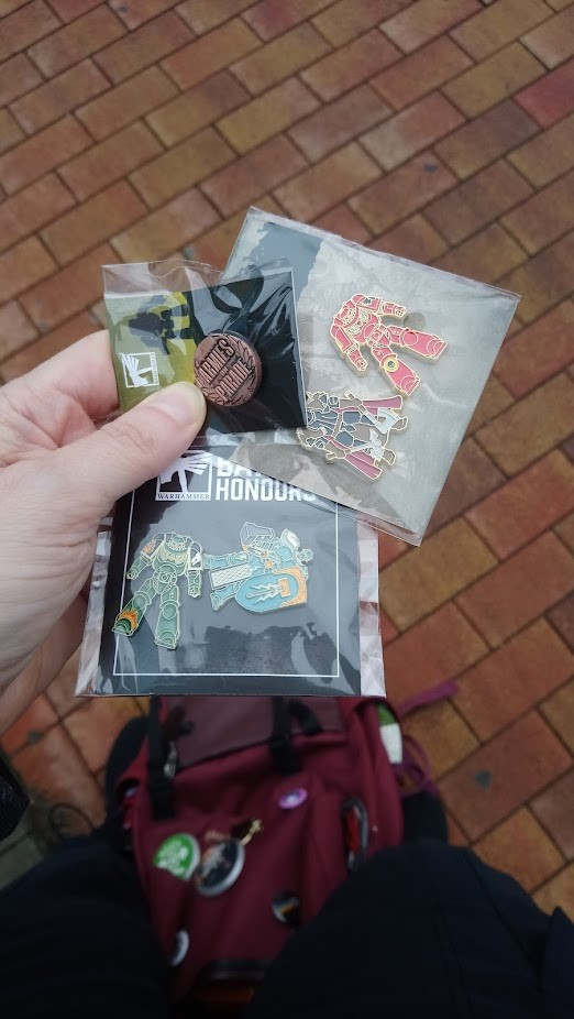 Photo of a hand holding three little plastic bags with pins. One with two Space Marines for Battle Honours, one with the Armies on Parade participation pin and one with the Marines/AoS-pin for Store Birthday.