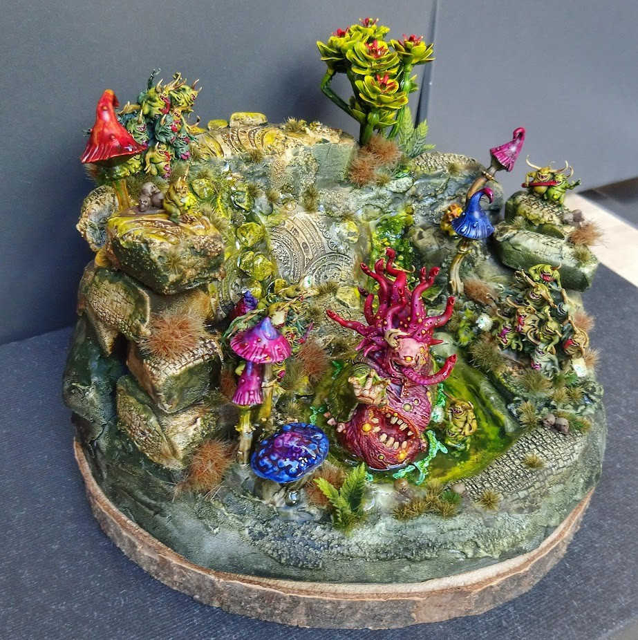 Photo of a little diorama. A small ruin landscape with a slimy waterfall and lots of damp mushrooms and mossy plants. A Beast of Nurgle is taking a bath in a pool and is getting washed (or slimed) by a lot of Nurglings with little sponges.