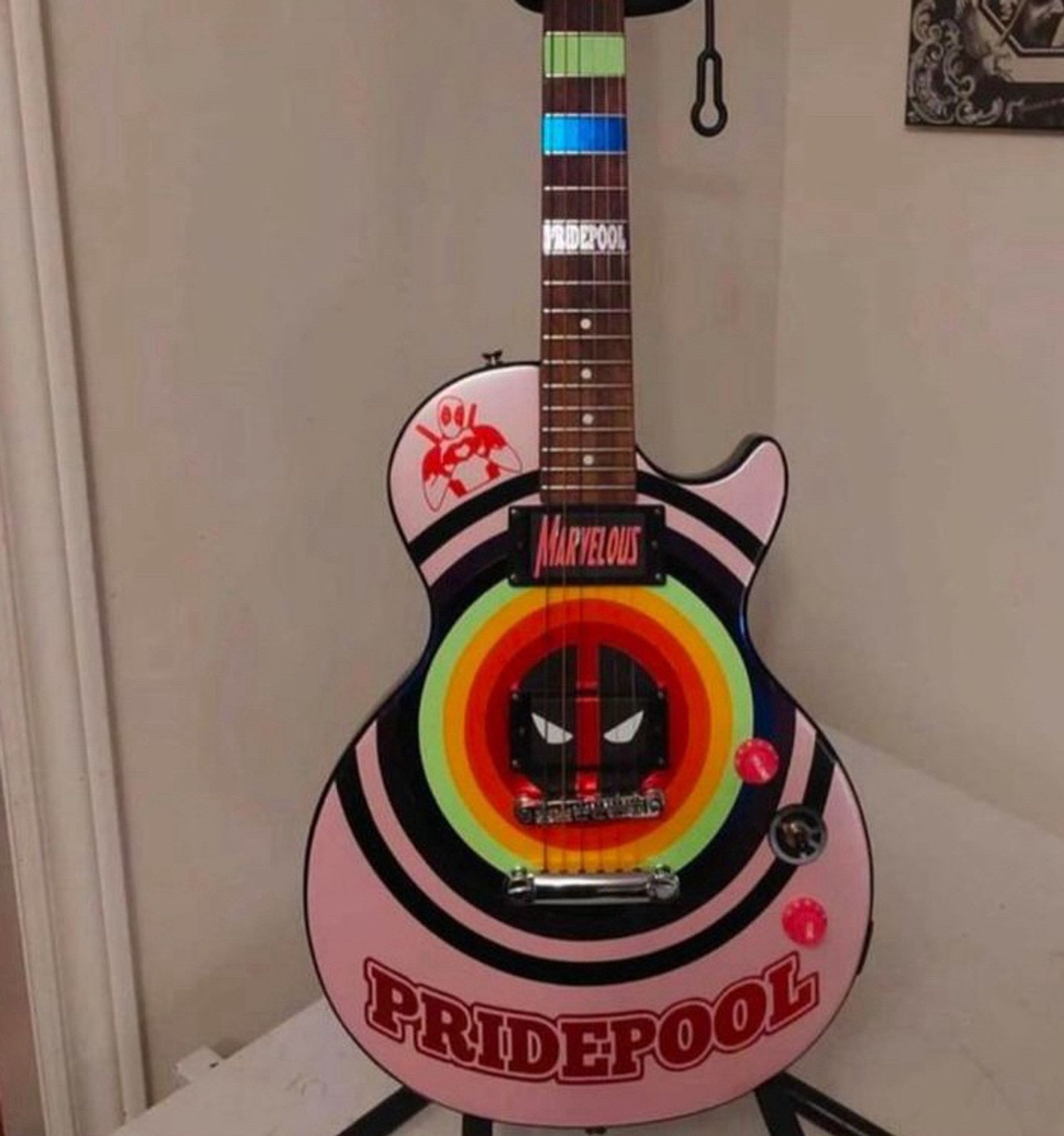 A pink guitar with a Les Paul shaped body; it has a bullseye pattern on the body that starts black, moves to rainbow, and ends in the Deadpool logo. At the bottom there’s text that says PRIDEPOOL