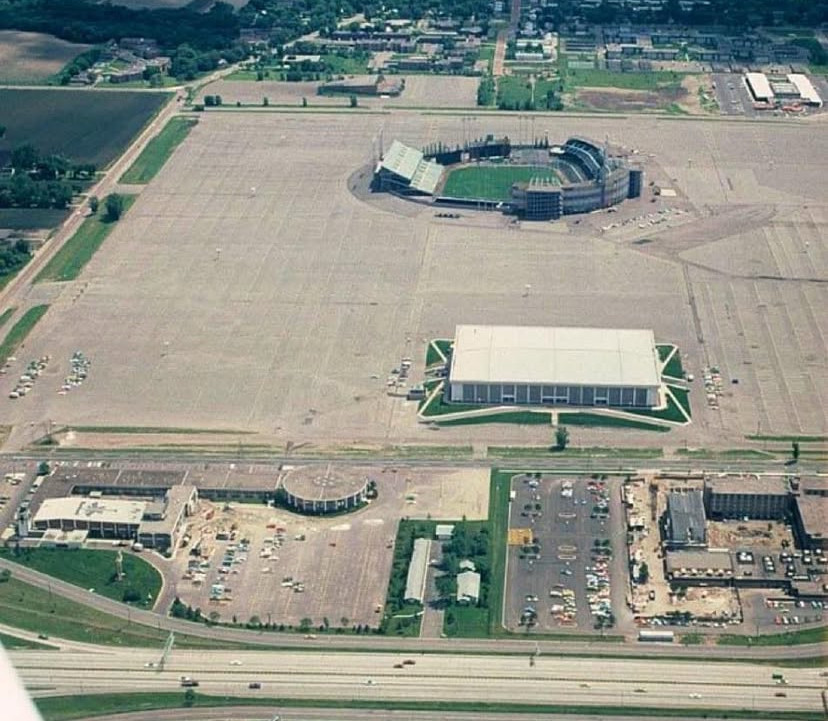 A stadium and a boxy building embedded in a mammoth parking lot; outside that, farmland