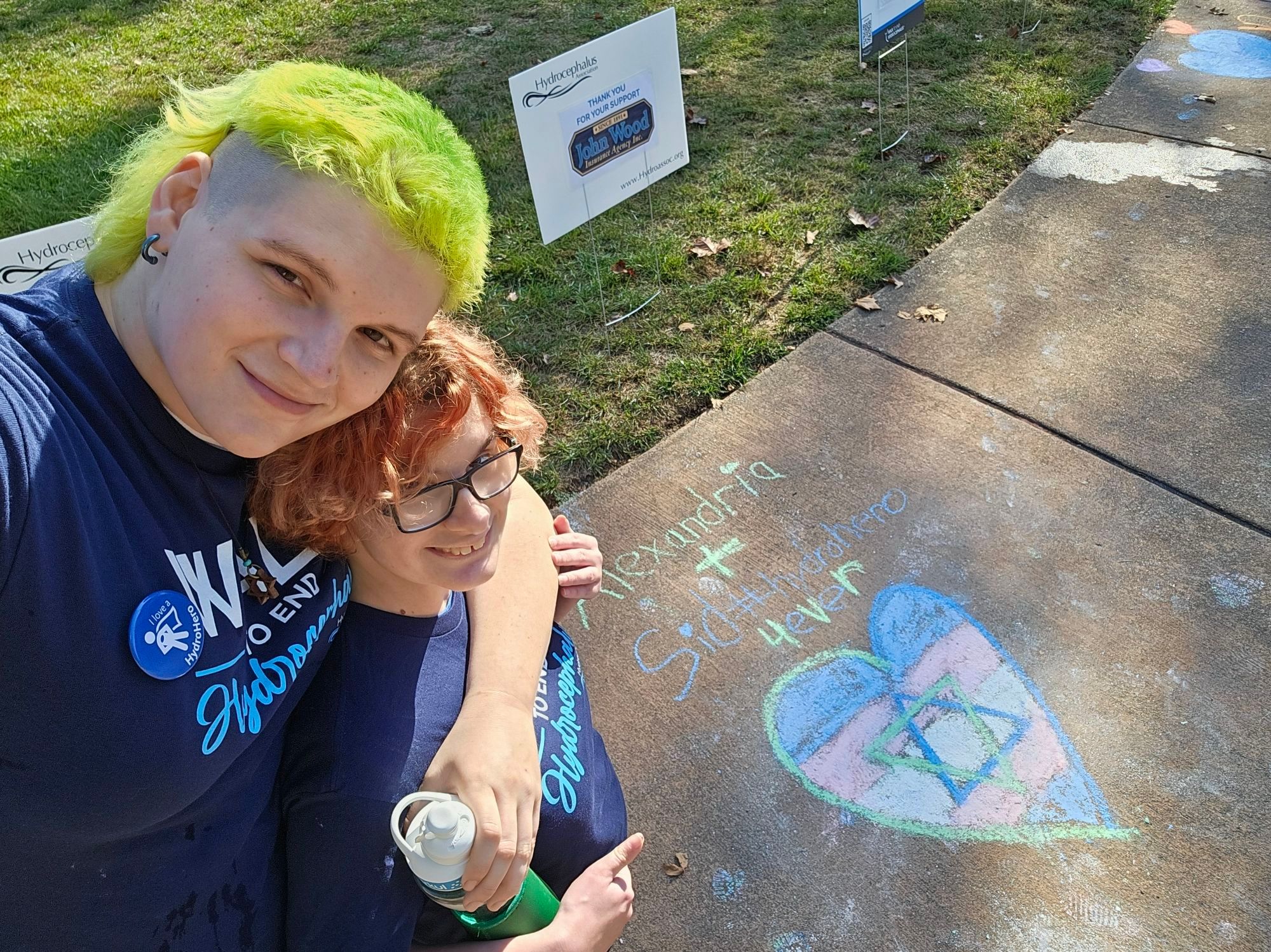 Me and my fiance, Sid standing next to a chalk drawing that says 'Alexandria + Sid #hydrohero 4ever' with a heart containing a trans pride flag and a magen david