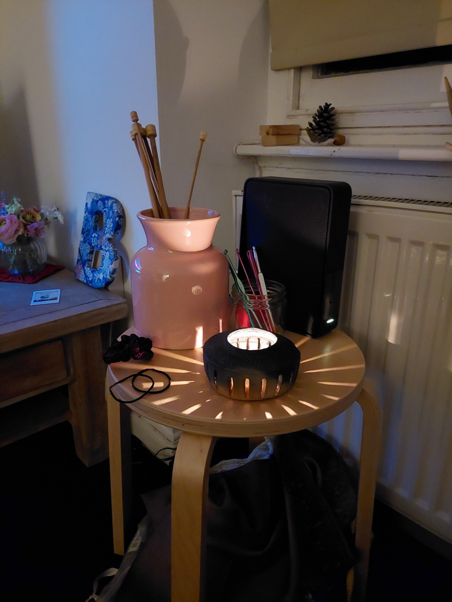 a photo of a replica bronze age pottery cup, which has a lit tealight candle inside. it has tall sides with vertical slices cut through at regular intervals so the light spills out in a sunburst and i LOVE IT. the cup is sitting on a little wooden side table in my living room, alongside my crochet hooks and knitting needles and internet  router. the table is in front of my window, you can see a bit of the windowsill and there's a pine cone on there that i picked up on a walk with my friend. the photo is warm vibes and brings me joy i hope you like it too xx