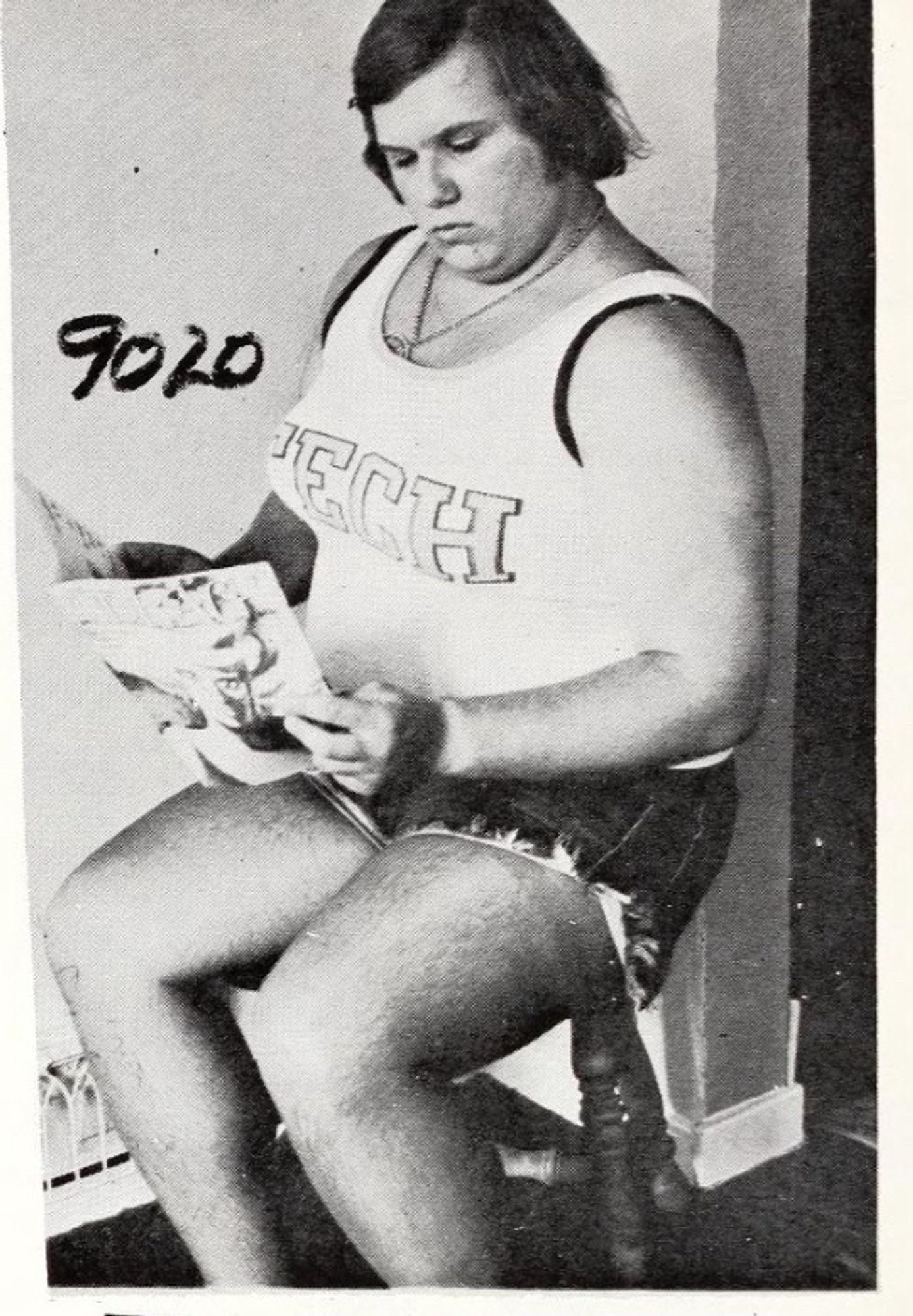 Black and white photo of a short-haired trans woman wearing a white tank top and cut off shorts, sitting on a stool reading a magazine.