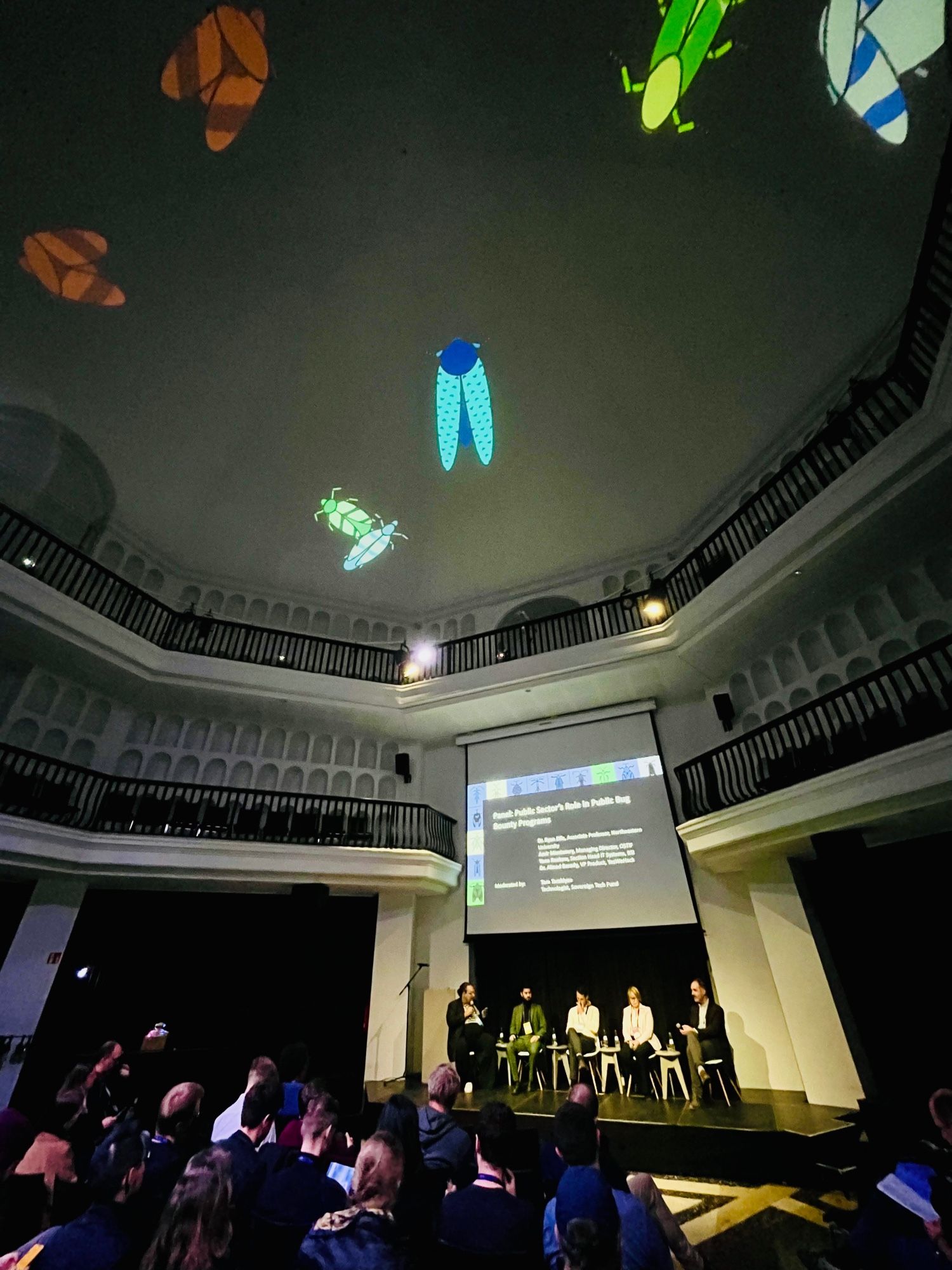 Wide angle view of panel on stage from left to right: Tara Tarakiyee, Amir Montazery, Dr. Aïmad Berady, Yona Raekow, and Dr. Ryan Ellis. Different colored generative art beetles are projected on the ceiling.