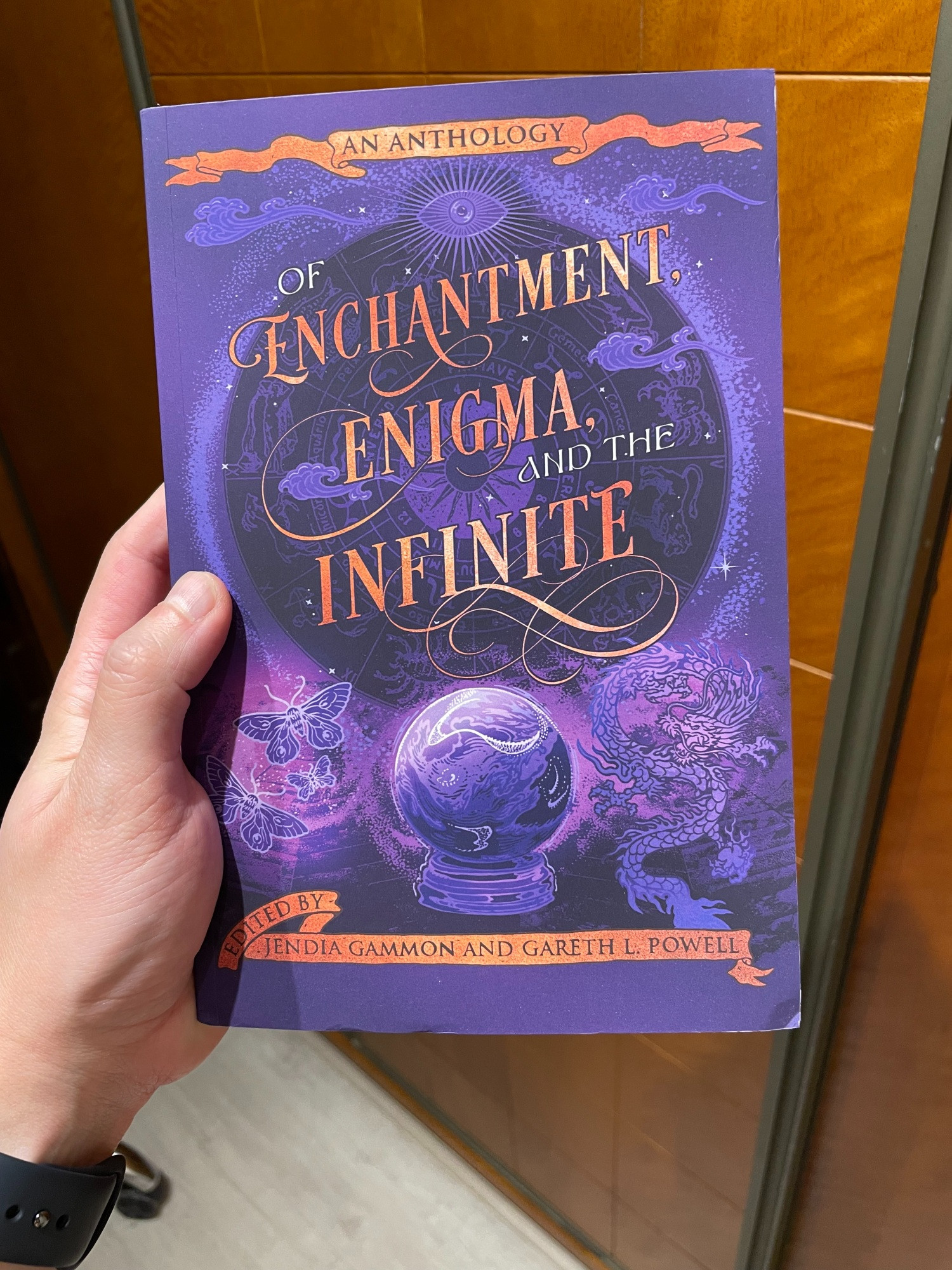 A white hand holding a book cover. The book is called "Of enchantment, enigma, and the infinite, edited by Jendia Gammon and Gareth L Powell." It features butterflies, a dragon, and a crystal ball in the center with some other fantastic motifs all around.