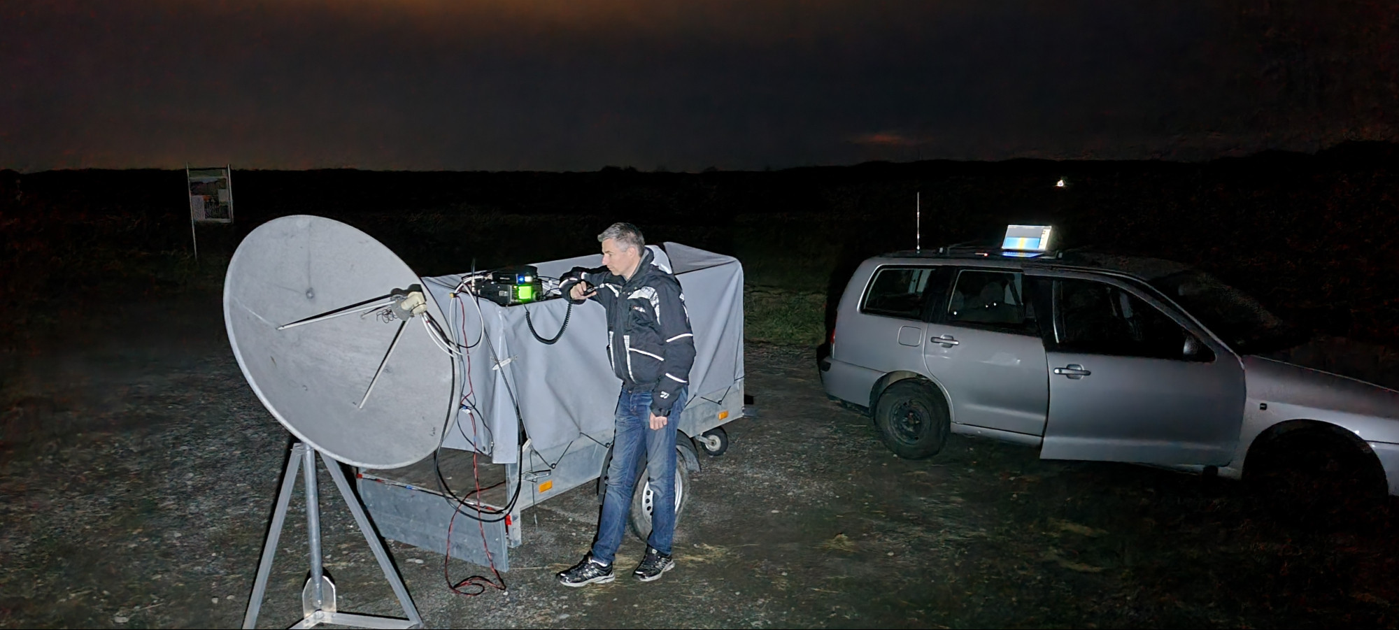 Ein Parkstreifen vor einem Feld. Es ist dunkel. Im Hintergrund steht ein Auto, darauf steht ein Notebook und leuchtet. Im Vordergrund ein kleiner Anhänger mit Plane, da drauf steht ein Funkgerät. Vor dem Anhänger steht ein 1,2m Spiegel auf Stativ. Ein DH8GHH steht am Funkgerät und tut so als führt er ein QSO.