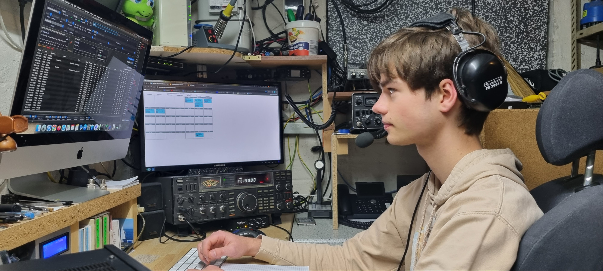 Konrad sitzt vor einer Funkstation, trägt ein Headset und schaut auf einen Monitor, auf dem das Logbuchprogramm zu sehen ist. Im Hintergrund steht ein Kurzwellentranceiver und darüber ein weiterer Monitor mit dem YOTA-Monat-Dezember Kalender