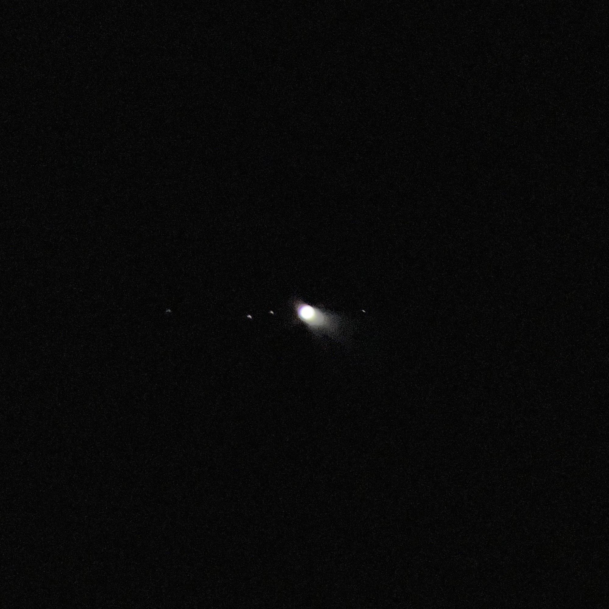 A blown out and somewhat fuzzy black and white image of a tiny Jupiter and even tinier moons. Taken with my iPhone held up to a small/cheap telescope eyepiece.