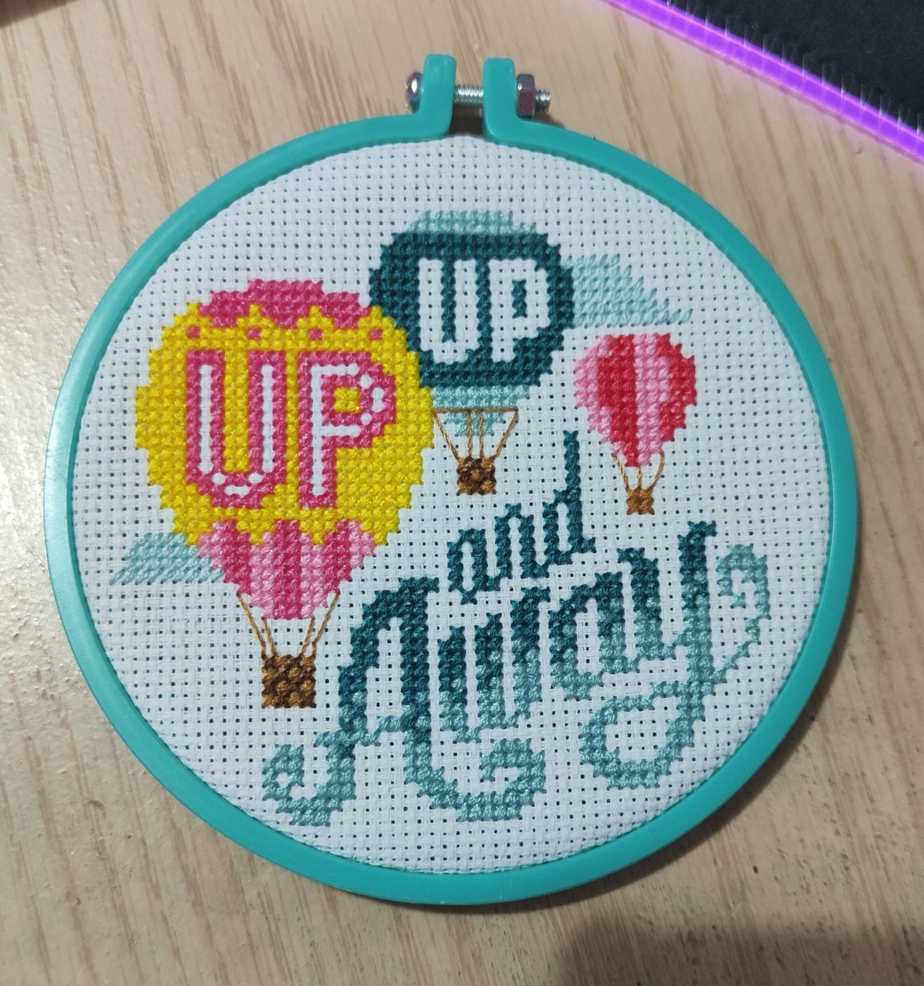 Small round cross stitch piece that says "up up and away" with three air balloons on it.