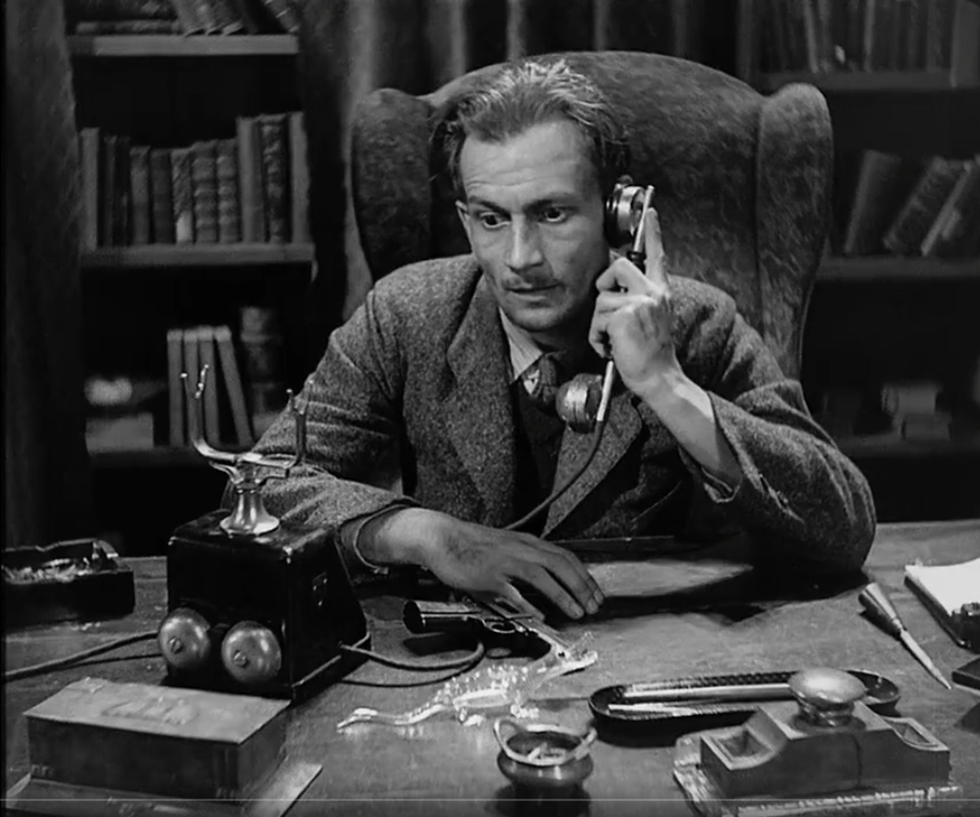 A still from the film, The Testament of Dr. Mabuse. A man is sitting at a desk and talking on the telephone, and in front of him is a little glass reptile.