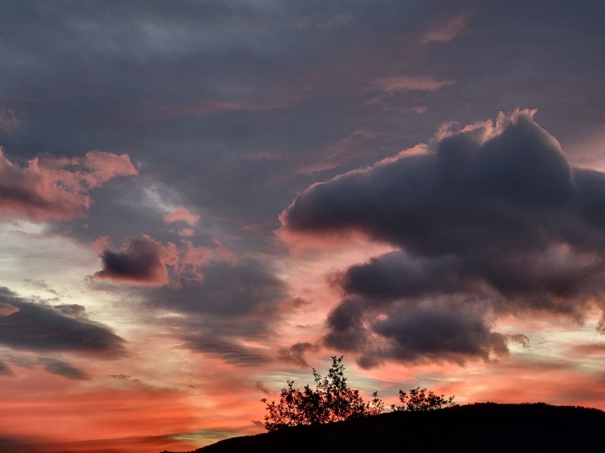 Nachdem die Sonne hinter einem dunkeln Hügel untergegangen ist, färbt sich der Horizont pfirsichrot. Darüber türmen sich aber dunkelgrau, bedrohliche Wolken.