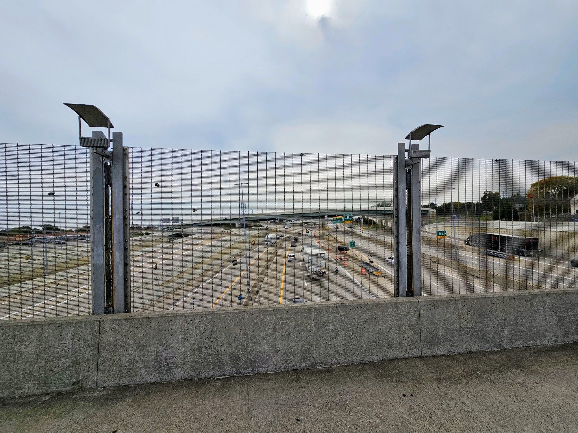 A photo from the center of the bridge, looking down at 10+ lanes of interchanging highway traffic. A few love locks are scattered along the fencing of the bridge.