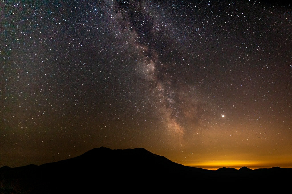 Mount Saint Helens in silhouette in front of the Milky way and yellow light pollution from Portland