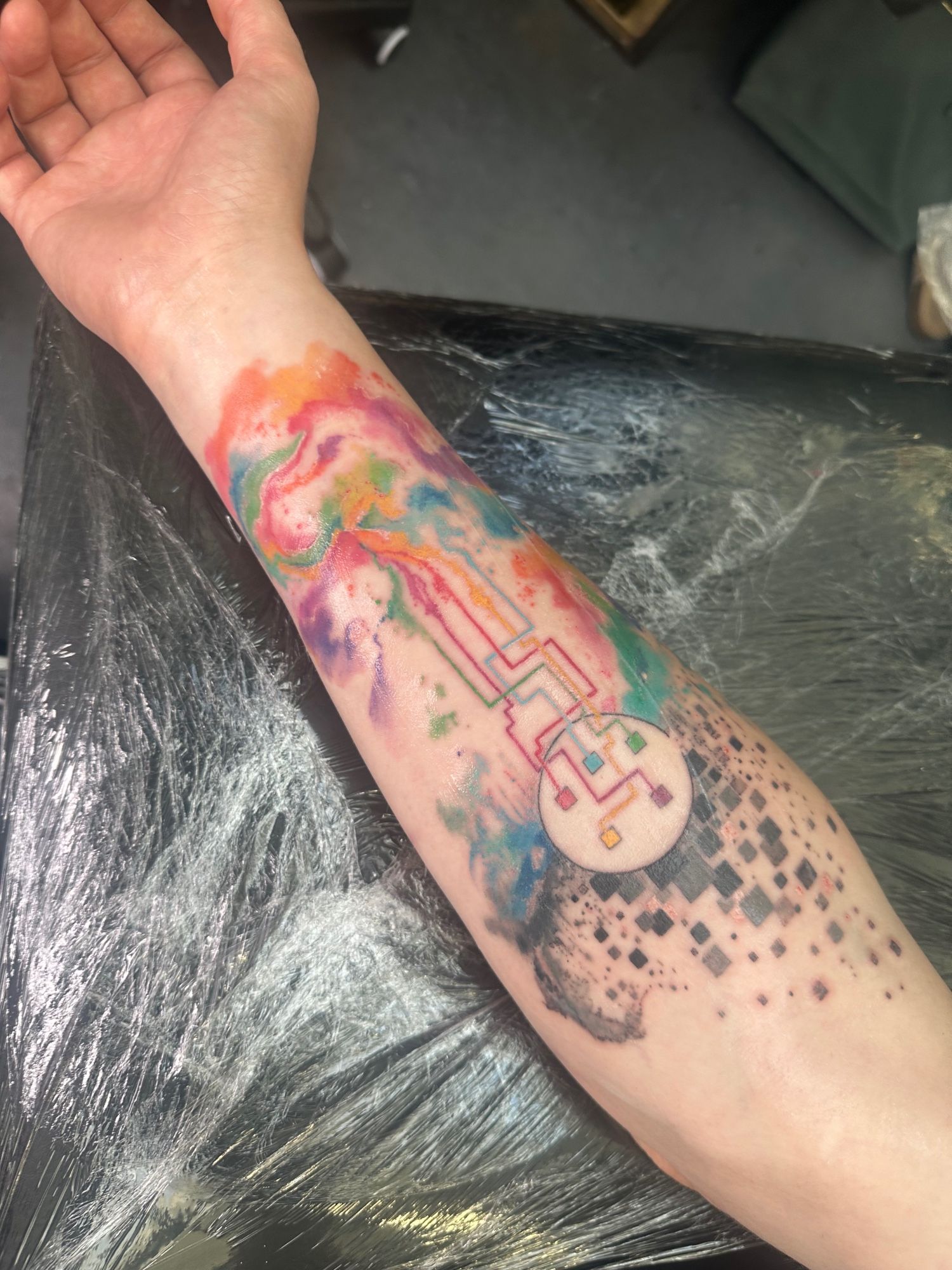 John’s arm with completed colourful tattoo that is somewhat abstract therefore hard to describe but goes from greyscale to multi colour going down the arm