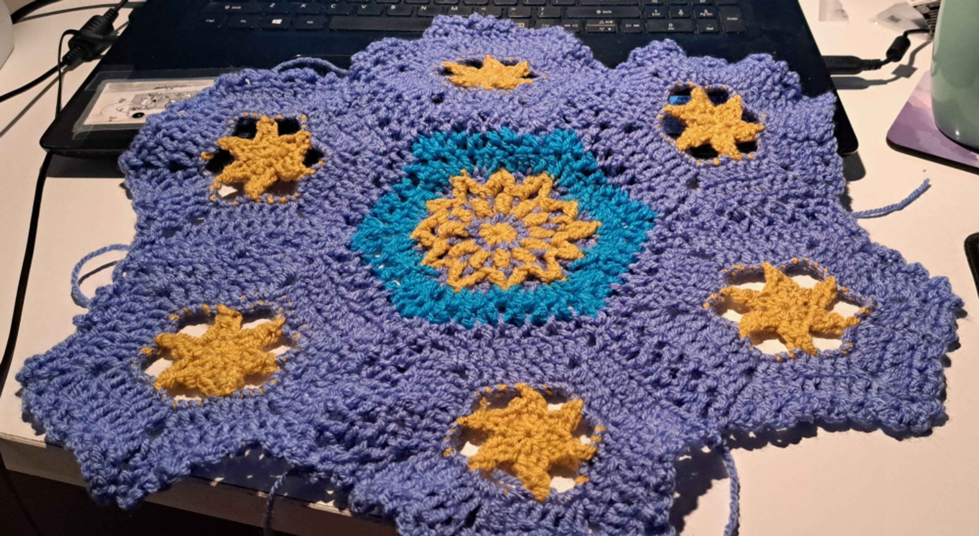 photo of a large crocheted hexagon consisting of seven smaller hexagons in various colors of blue, purple, and yellow. Each smaller hexagon has a yellow star or sun at the center.