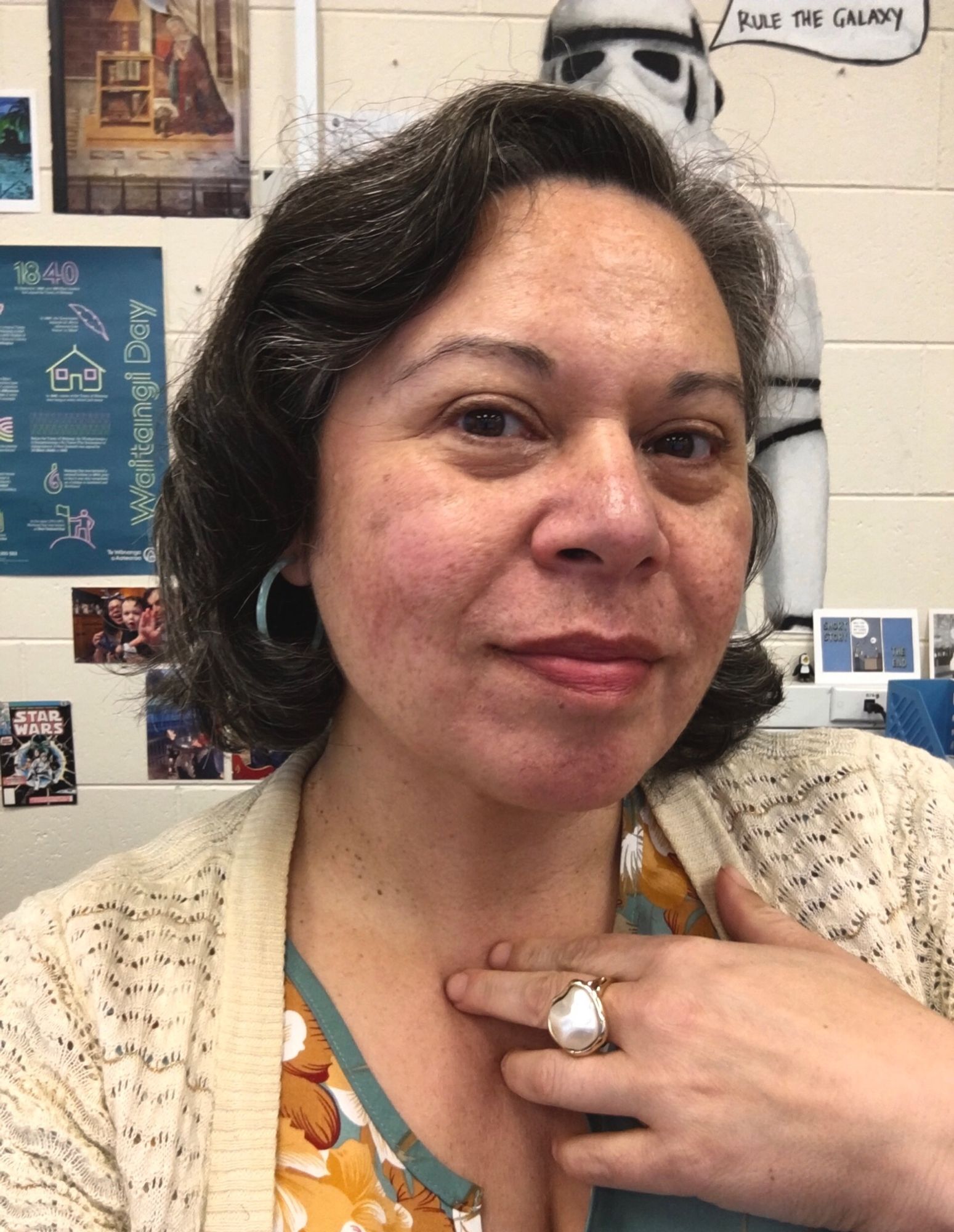 Head and shoulders selfie of a middle aged Māori woman. She has a large cocktail ring and green plastic hoop earrings on.