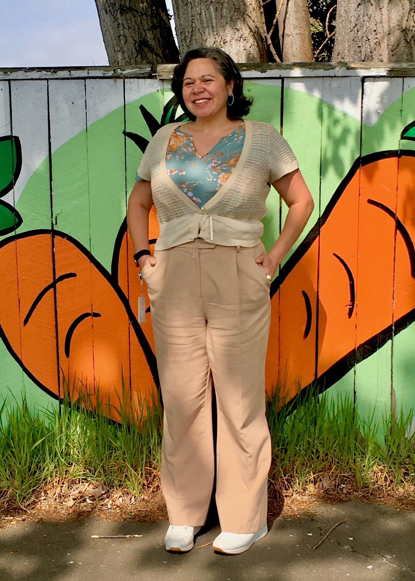 Full length photo of a middle aged Māori woman wearing tan wide legged trousers with white wedge sneakers. She has a cream short sleeved cardigan on over a green floral blouse.