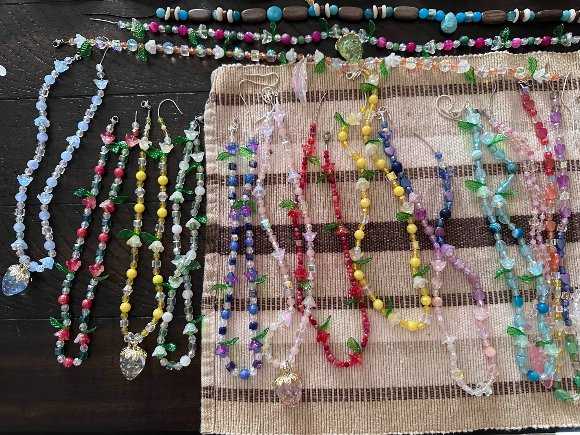 a ton of unfinished necklaces (ends haven't been put on them yet) in varying colors, all cute chunky beaded necklaces - some have strawberry charms, some have flower and leaf beads, some have both strawberry charms and flower/leaf beads - there is also one turquoise drop necklace with seashells and wooden and glass/acrylic beads on a black chain. Every other necklace minus the turquoise drop one is on a stainless steel chain.