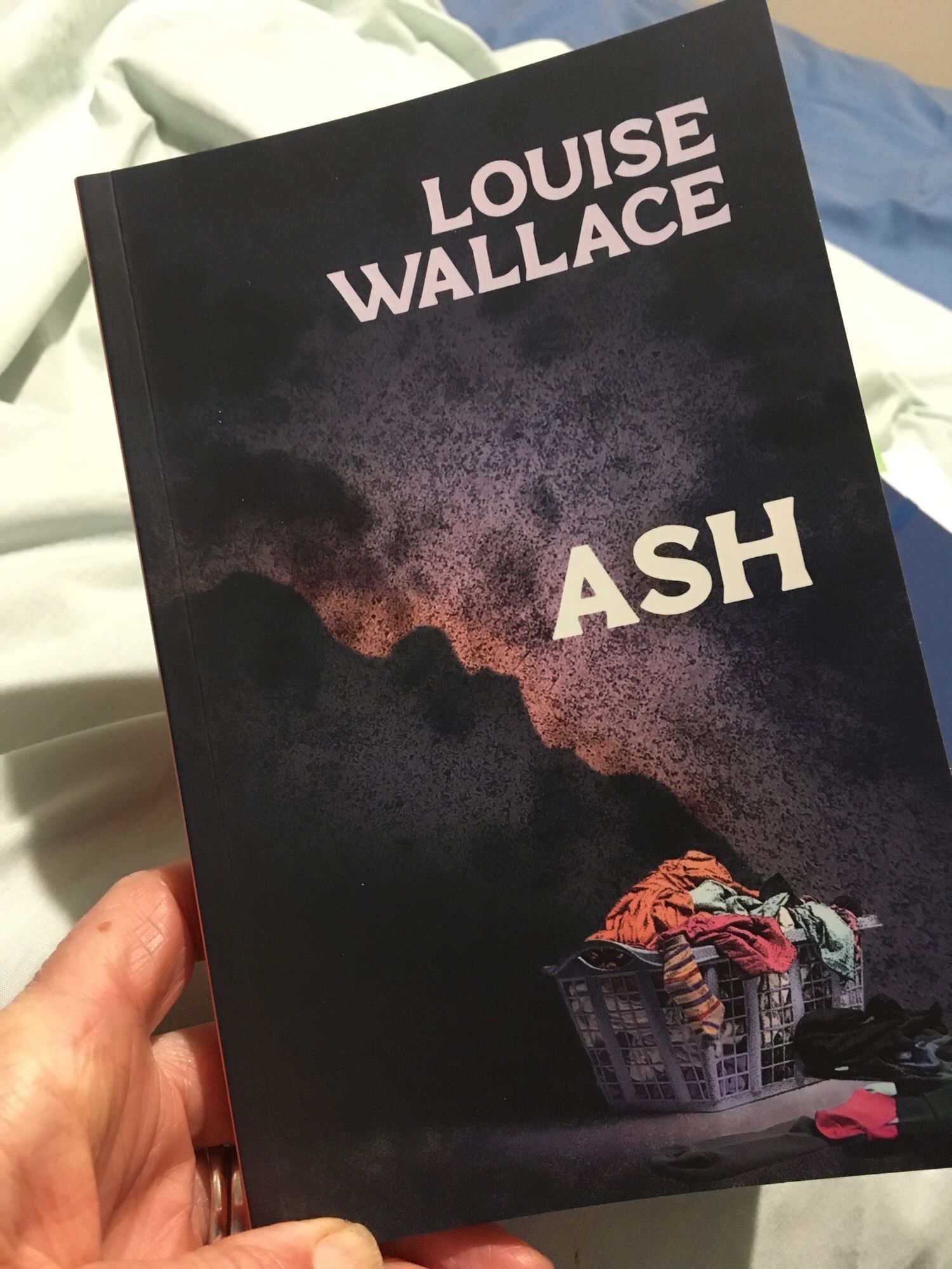 The cover of Louise Wallace’s book Ash; it shows a colourful, chaotic basket of laundry with a volcanic explosion in the distance.