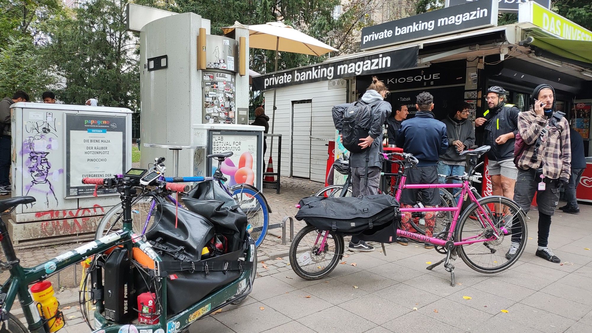 Transportfahrräder vor einem Kiosk und Leute, die sich unterhalten oder telefonieren. Im Hintergrund eine öffentliche Toilette und ein Schaltkasten, beide voll mit Aufklebern.