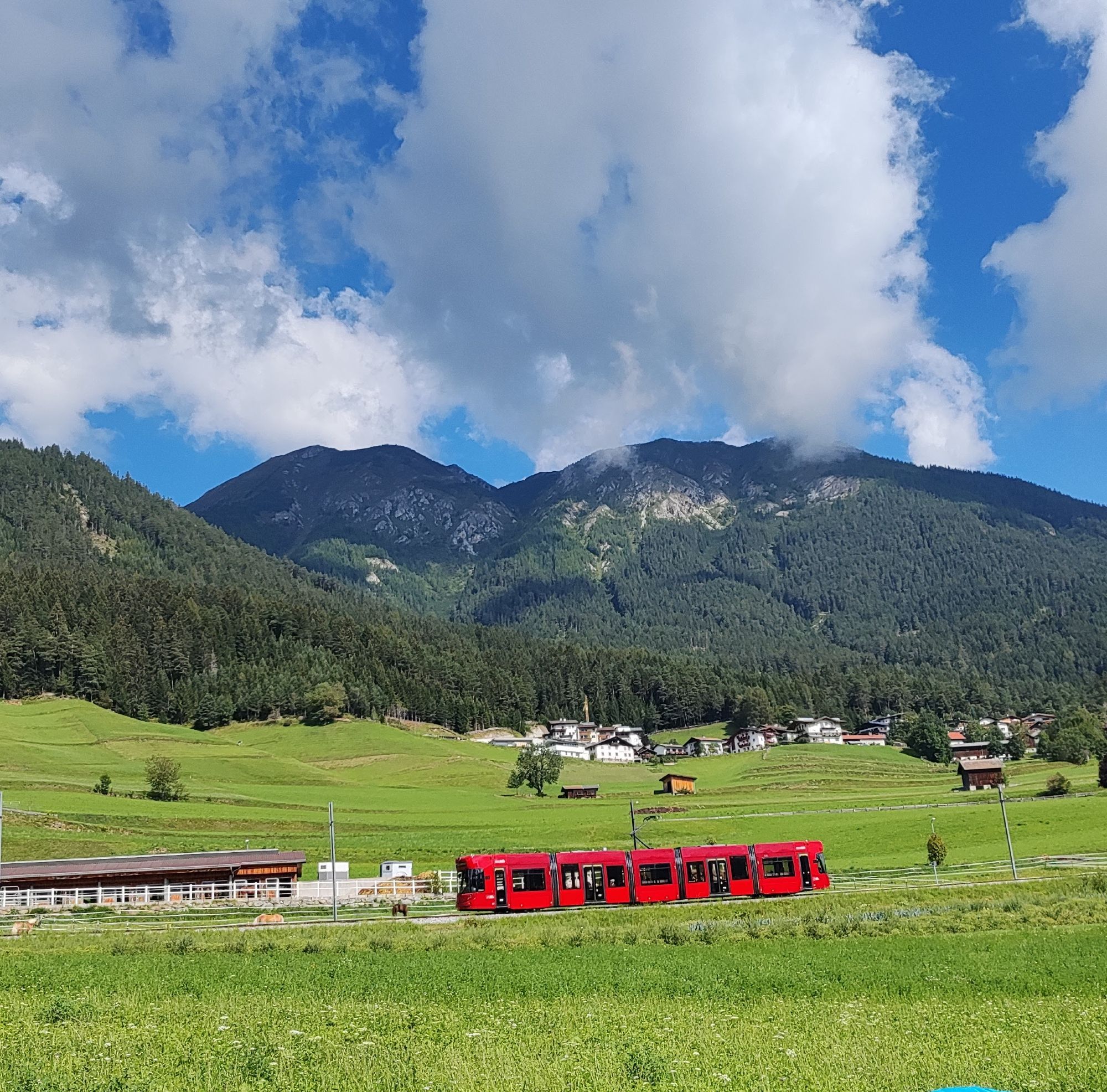 eine rote straßenbahn in grüner Wiese dahinter Berge und Himmel mit weißen Wolken