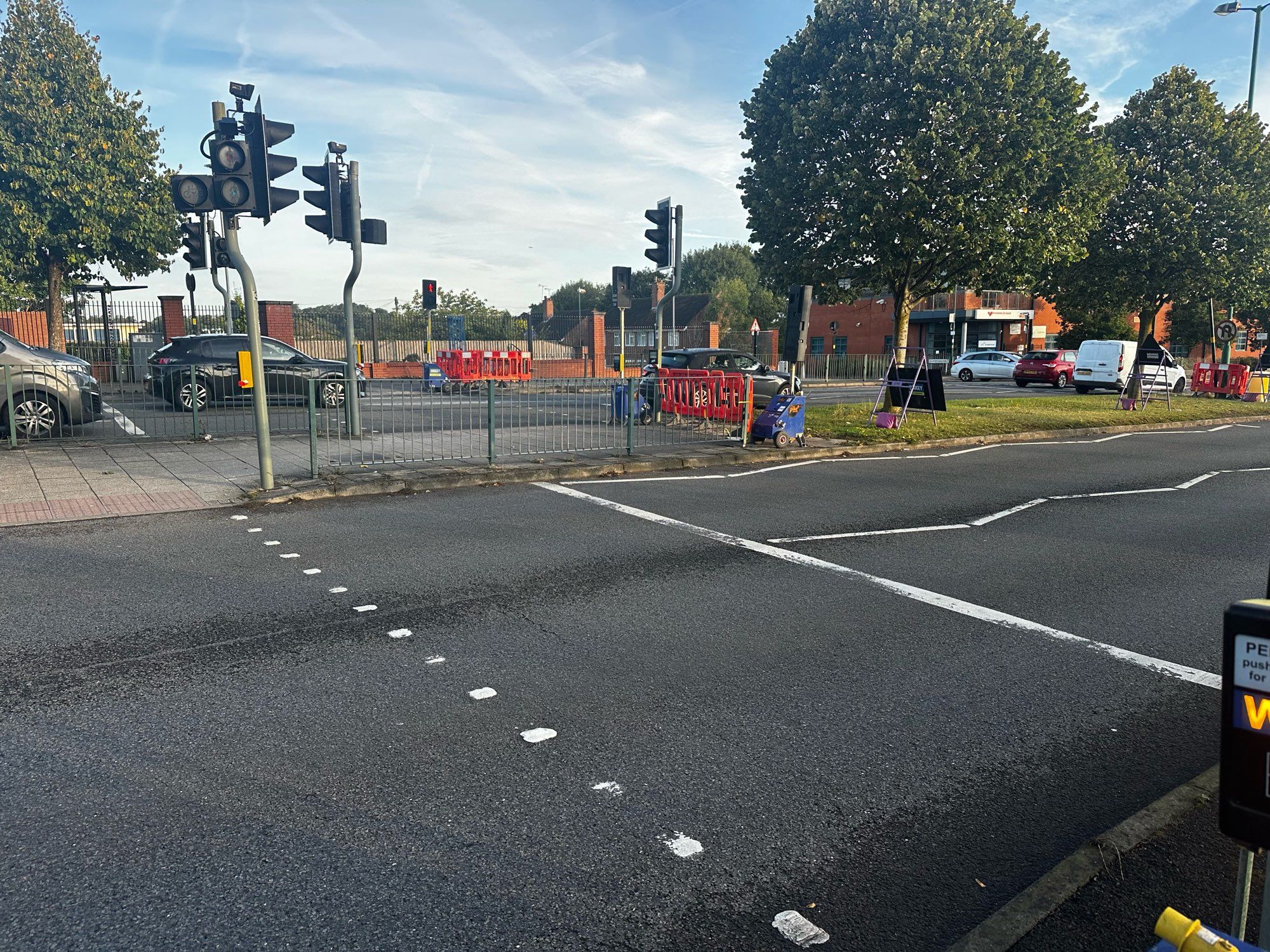 A dual carriageway local road. There are pedestrian crossings across both which are replaced by temporary lights. Metal railing is replaced with plastic temporary barriers. A lamppost has been knocked down. A signpost has been knocked down.