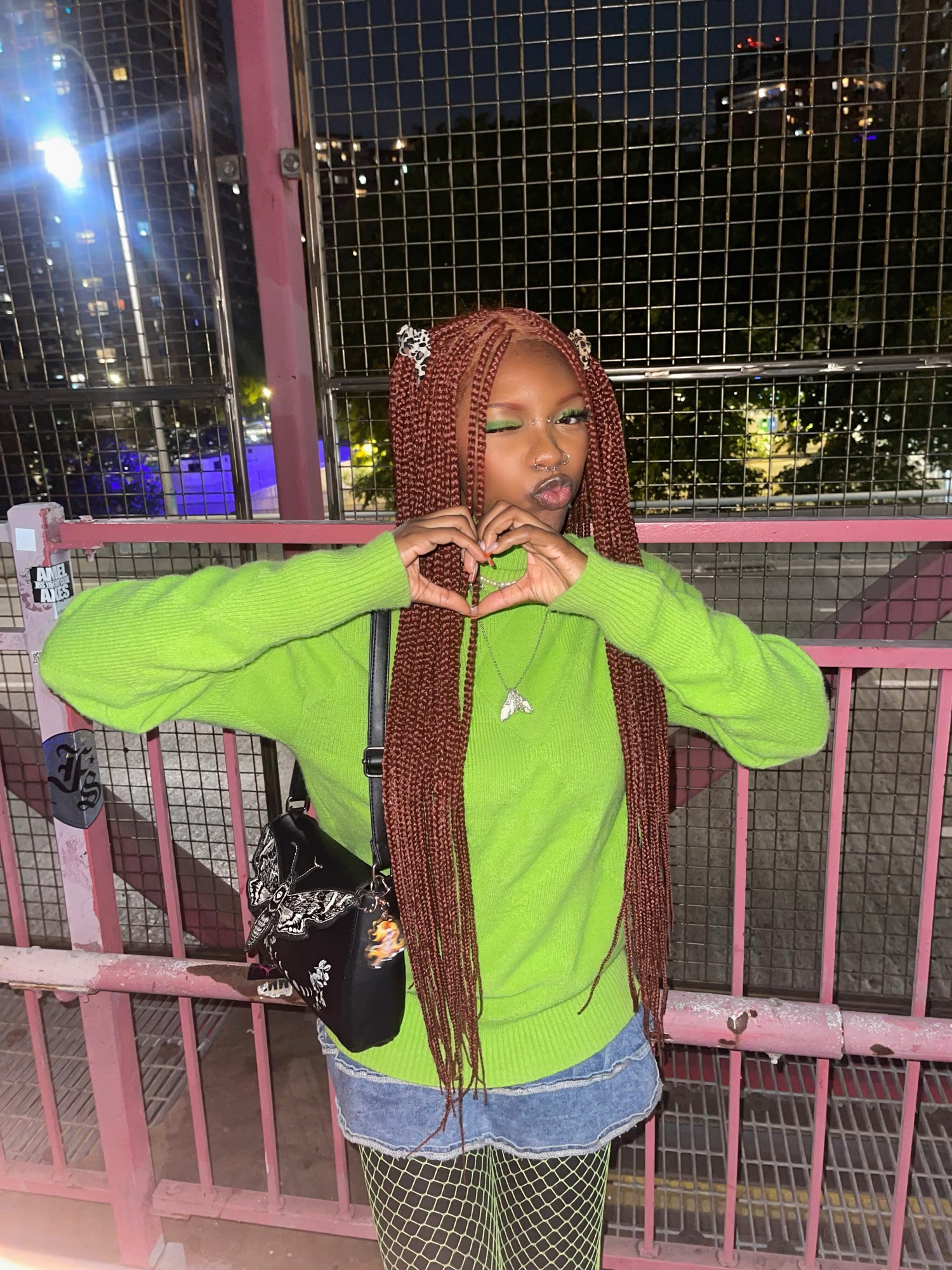 a fem presenting person in a oversized lime green sweater, ginger braids, green fishnet, and green eyeshadow. the person in the photo is making a kissy face while holding their hands in the shape of a heart.