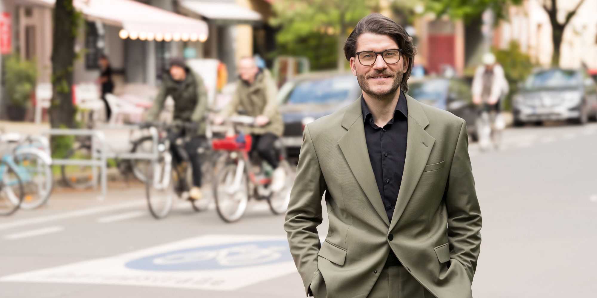 Ein Mann blickt frontal in die Kamera. Im Hintergrund ist eine Fahrradstraße mit zwei Radfahrenden zu sehen.