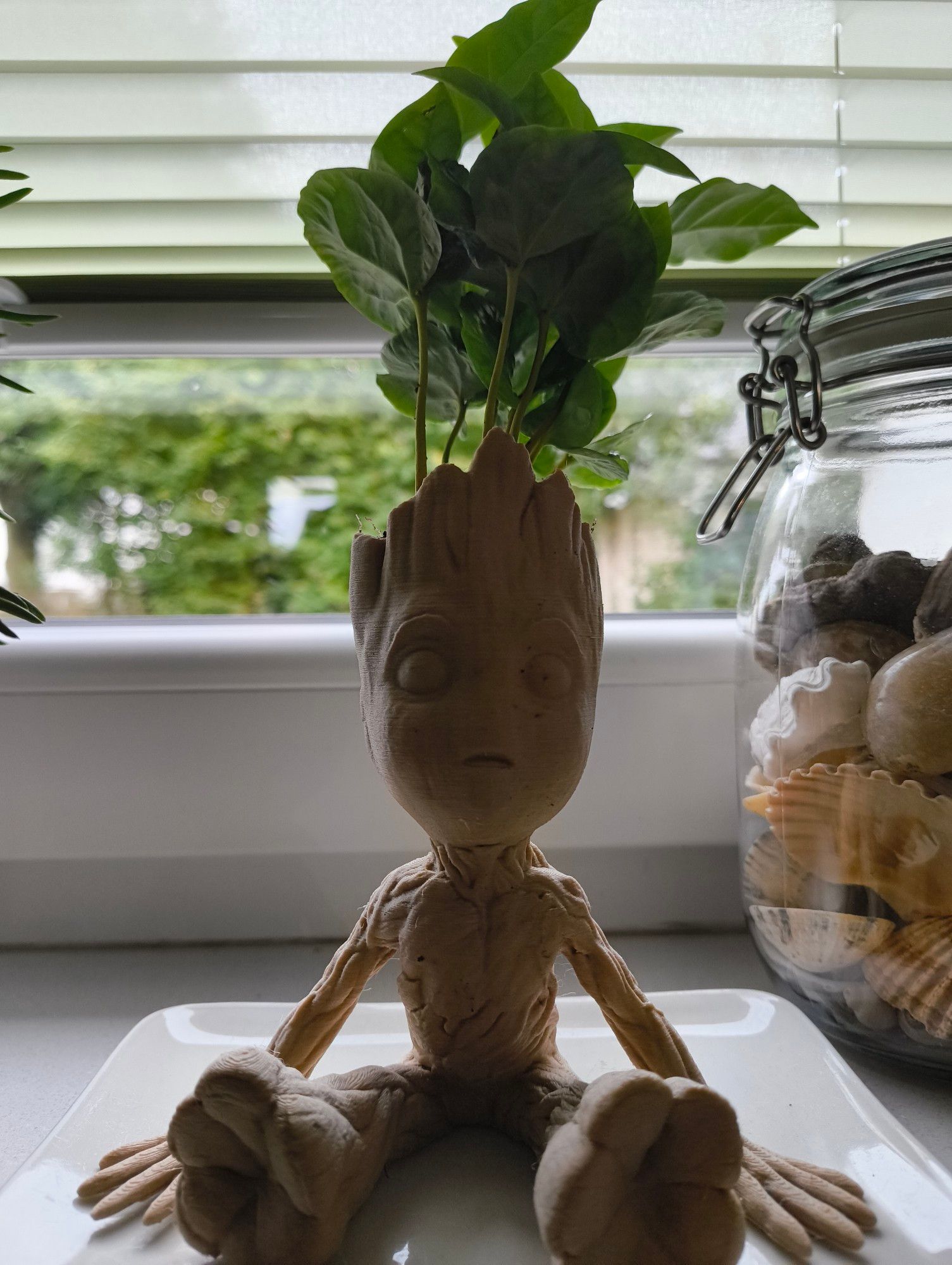 Ein sitzender Baby Groot, dessen Kopf als Planttrog dient. Eine grüne Pflanze "wächst" aus seinem Kopf. Die Figur ist 3D-gedruckt, mit einem Material, das zum Teil aus Holz besteht und deshalb hellbraun ist.