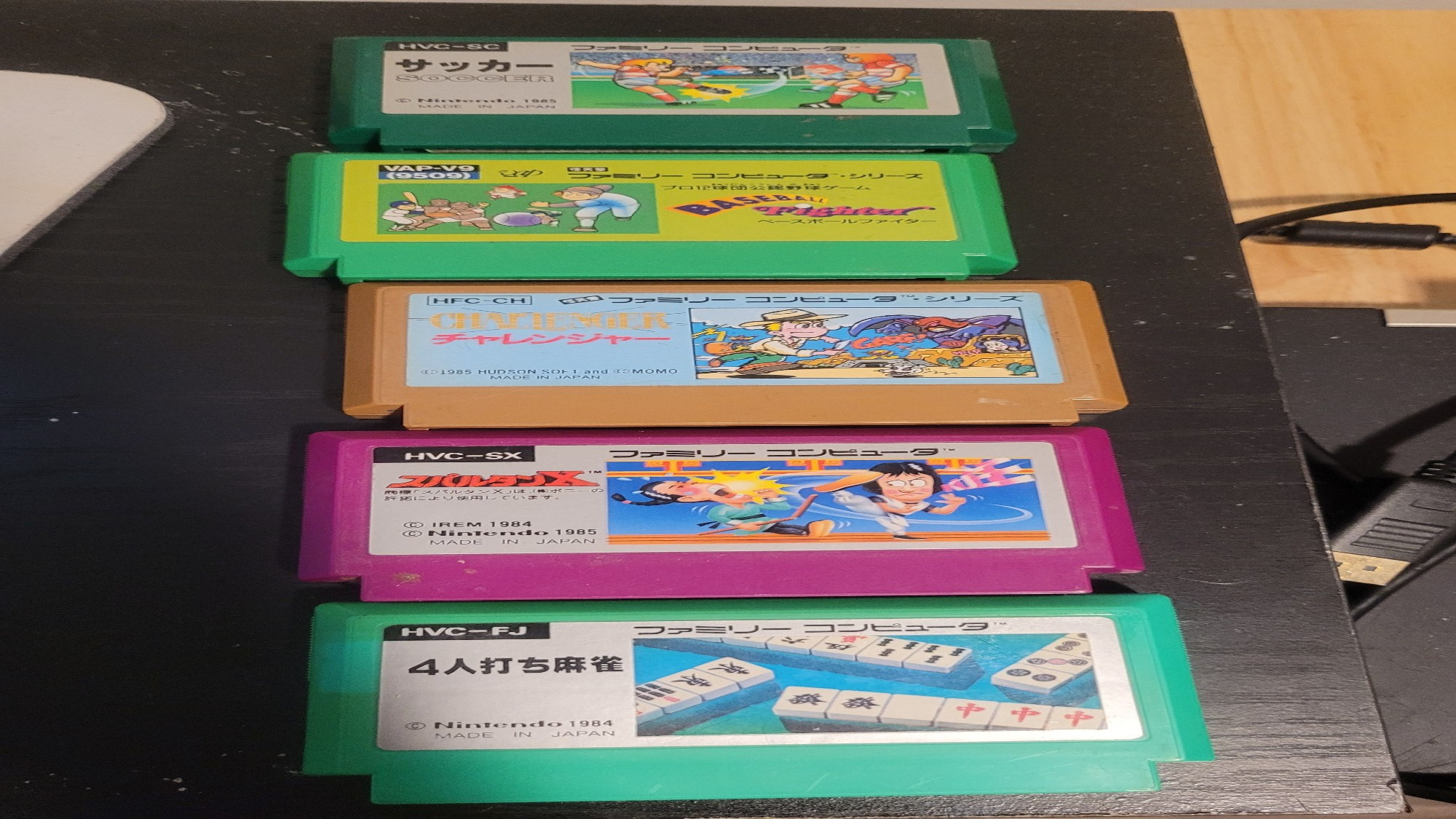 5 colorful famicom cartridges lined up vertically on a black desk. Among them are a soccer game, a baseball game, a game called Challenger, a game called Spartan X, and a Mahjongg game.