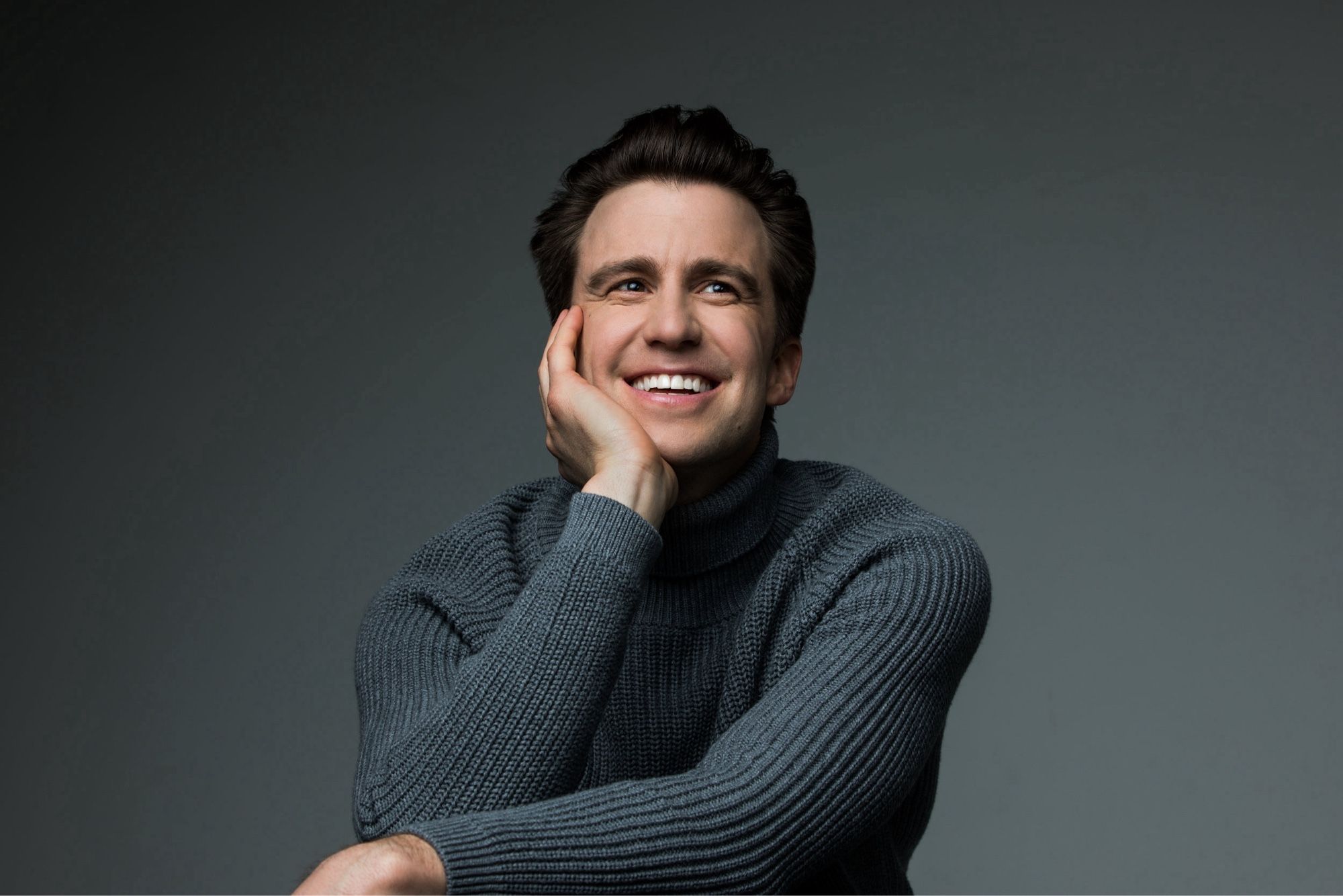 A portrait of the late actor Gavin Creel, smiling, in a cable knit sweater