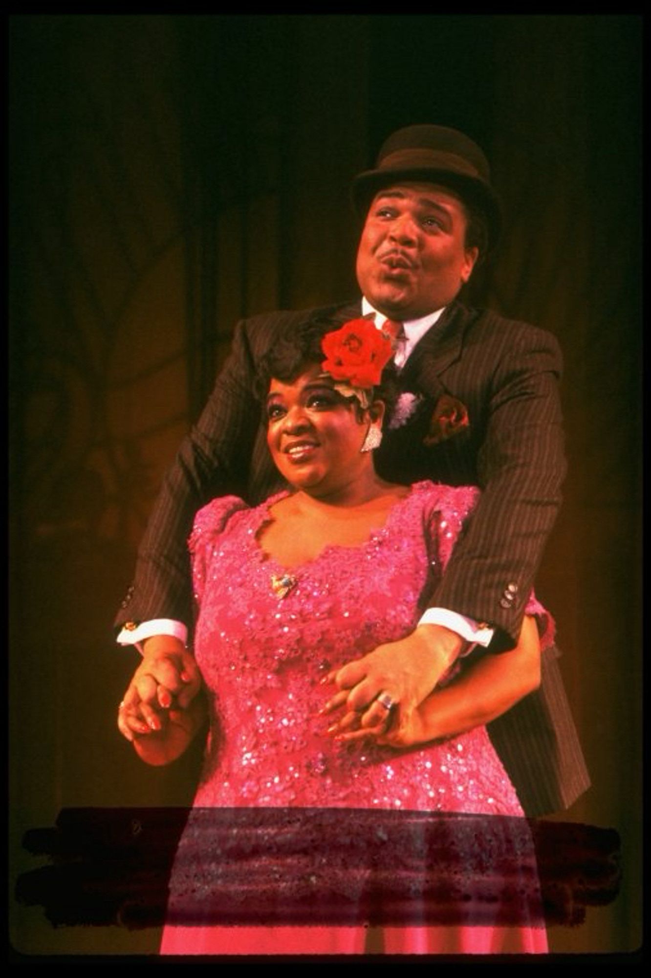 Ken Page in suit and bowler hat sings with his arms around Nell Carter, who wears a pink sequined gown and a red flower in her hair.