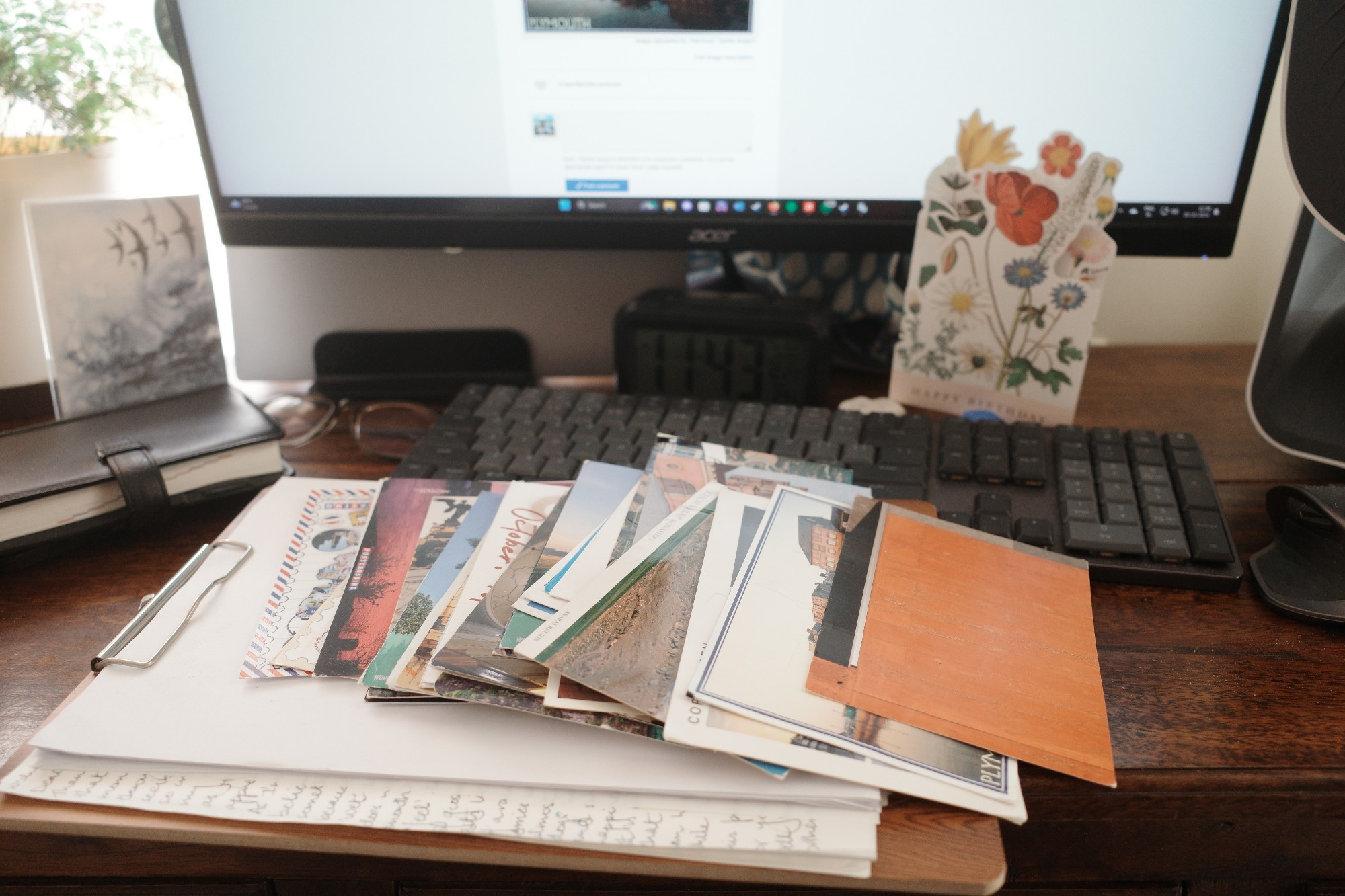 Pile of postcards from across the globe on my desk with a rabbit foot fern, framed postcard of a stormy sea, desktop computer and Hobonichi Techo journal in the background.