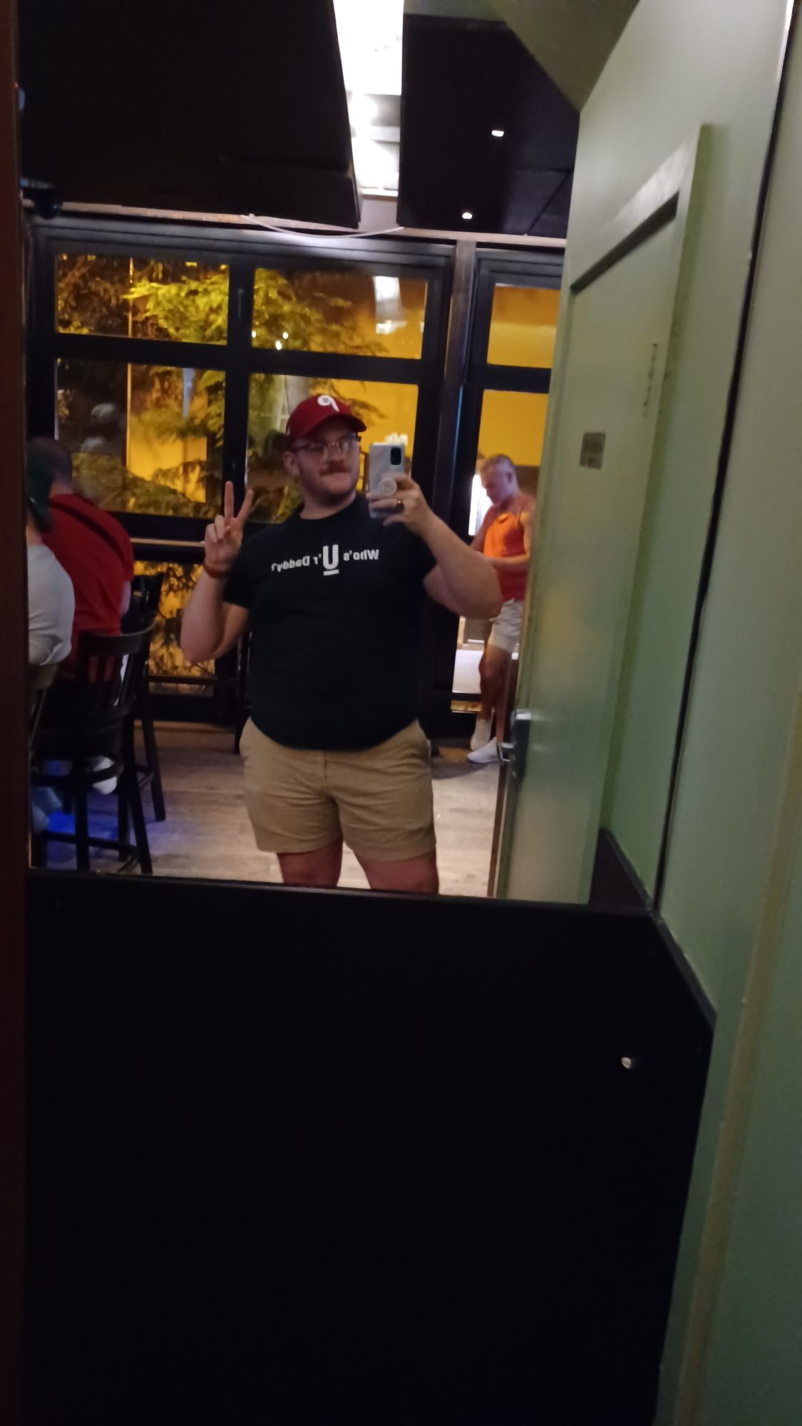 Mirror-selfie in a bar. I wear a Philadelphia Phillies cap, a black tee that says "Who's Ur Daddy", and short Khaki shorts. I am posed with my phone in one hand and the other forming a peace sign.