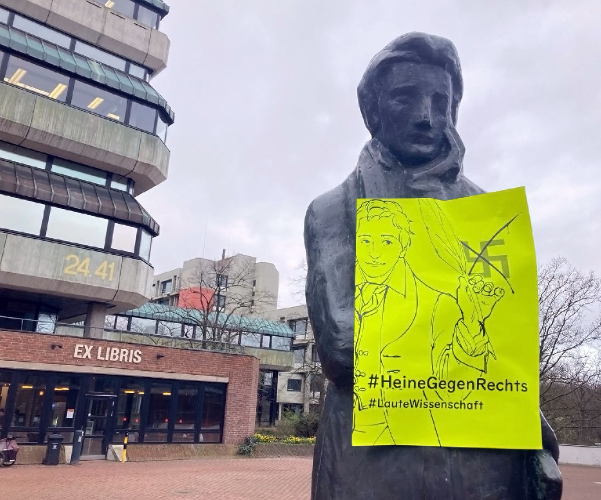 Ein Foto vom Campus der HHU. Im Vordergrund ist eine Statue von Heinrich Heine zu sehen, an welche ein neongelbes Plakat geklebt ist. Auf dem Plakat ist ein gezeichneter Heine, der ein Hakenkreuz mit seiner Schreibfeder durchgestrichen hat. Dazu die beiden Hashtags HeineGegenRechts und LauteWissenschaft. Im Hintergrund sind Teile anderer Uni-Gebäude zu sehen.