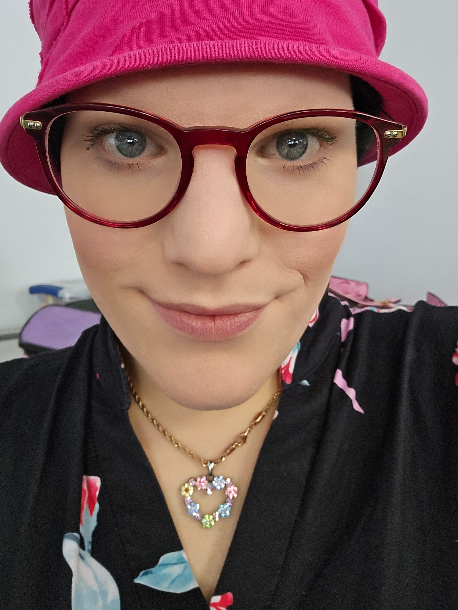 A close up photo of woman with a pink hat, ruby round glasses and a black floral top, with a necklace that has a heart shaped pendant made out of tiny flowers.