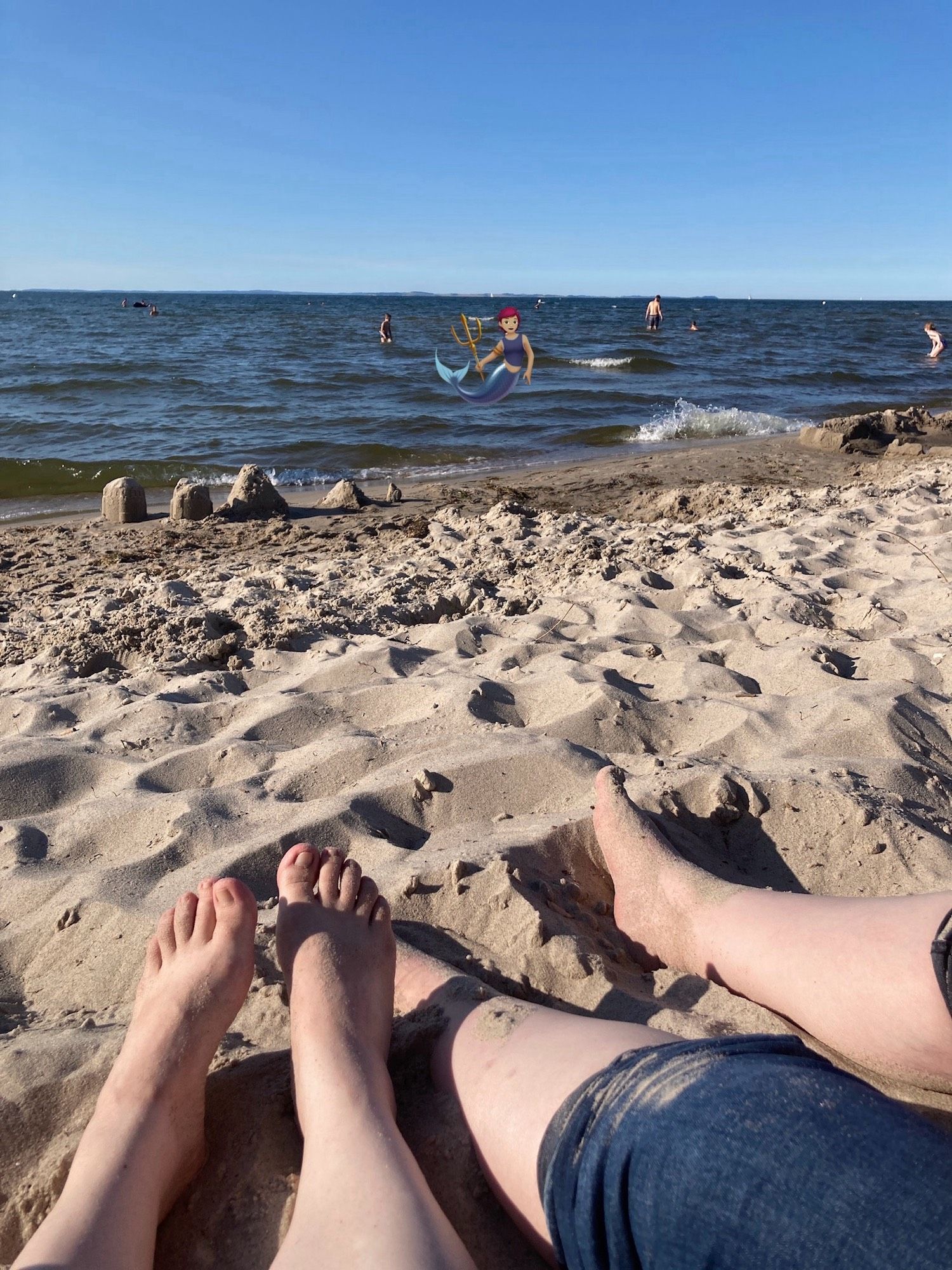 Our feet and legs on a sandy beach, trousers  rolled up. Sunny weather, people in the water.