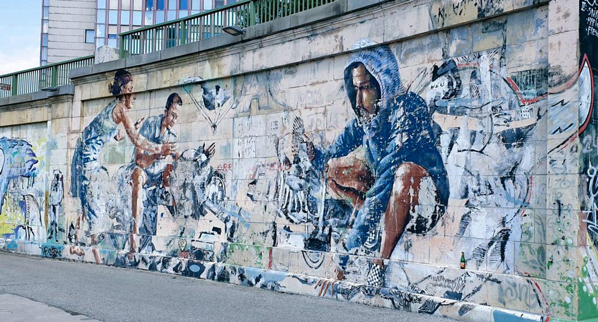 Graffiti on a slightly curved, light-coloured stone wall. The main character is a man in a blue hoodie, sitting and with his hood pulled over his head.
To the left of this figure are a man and a woman, looking as if they are trying to protect something or someone with their arms stretched out in front of them. The colours of the mural are very faded and some of them have already come off, so the artwork is no longer clearly recognisable.