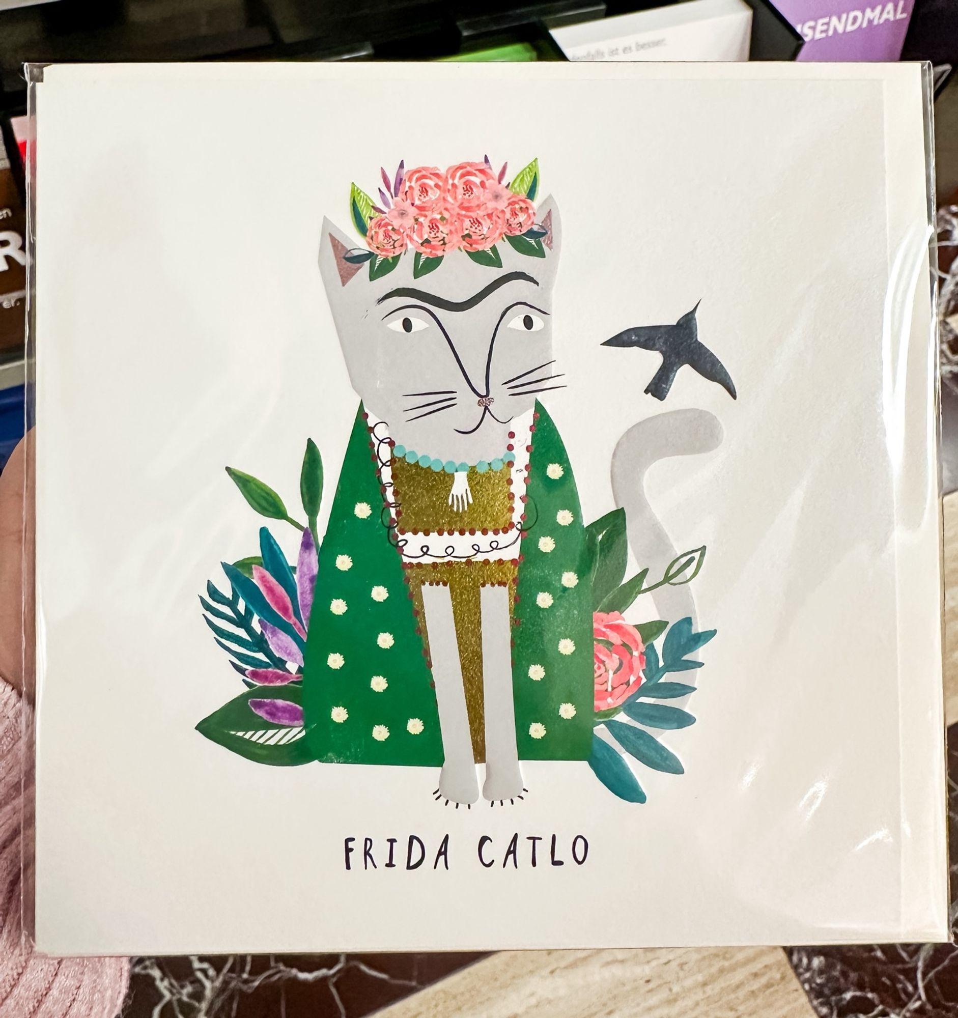 A sujet on a card showing a cat being dressed and looking kind of Frida Kahlo with flowers and leaves around the cat and on the cat‘s head. Below the cats is written Frida Catlo