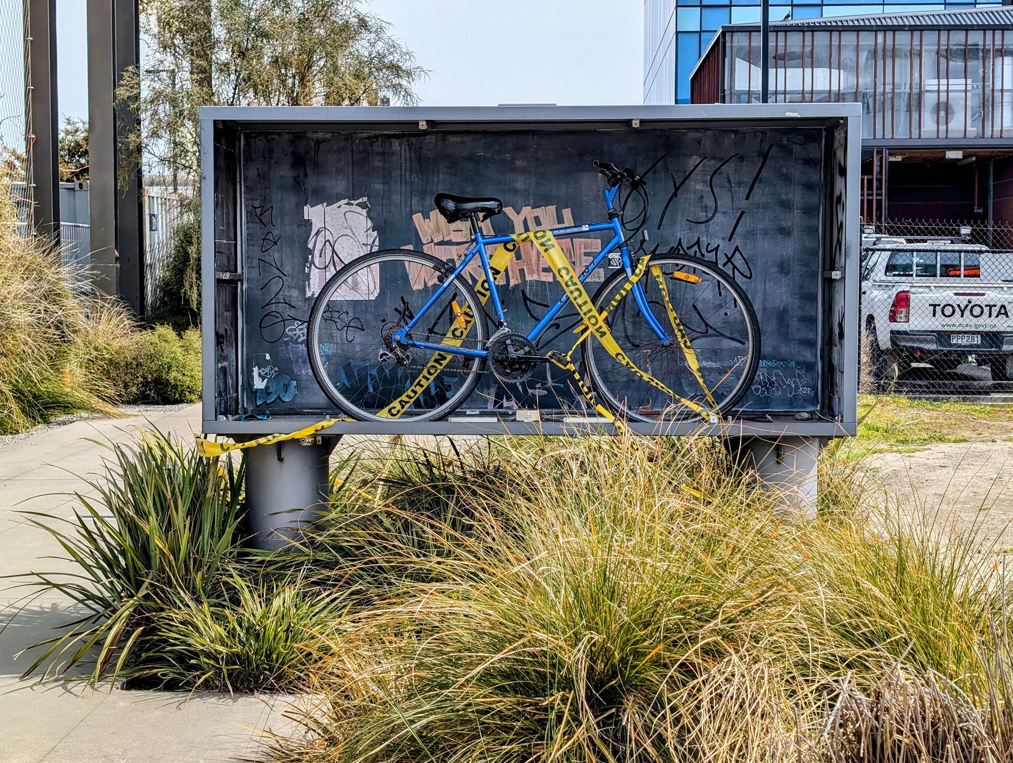 A metal rectangular box elevated on short pillars which used to contain the illuminated text "Wish you were here" until it was smashed long ago. The box now contains a blue bike with a bright yellow caution tape draped across. The back of the metal casing is covered in grafitti, including (quite poetically) the text "Wish you were here". In the foreground is a grassy flaxy rain garden. In the background are a building, a ute, and the concrete path of the Christchurch South Frame.