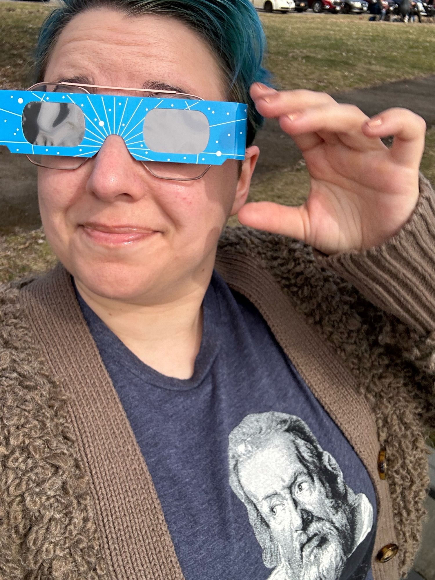 Blair, a white woman with short blue hair, is wearing a blue t-shirt with Galileo on it and a brown fuzzy sweater. She’s got eclipse glasses over her regular glasses and is gesturing towards them.