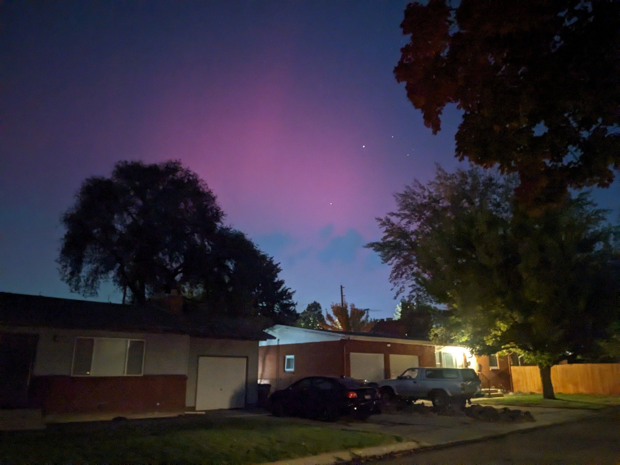 The pink and purple hues of aurora borealis, punctuated by stars, in the night sky above ranch-style houses and trees.