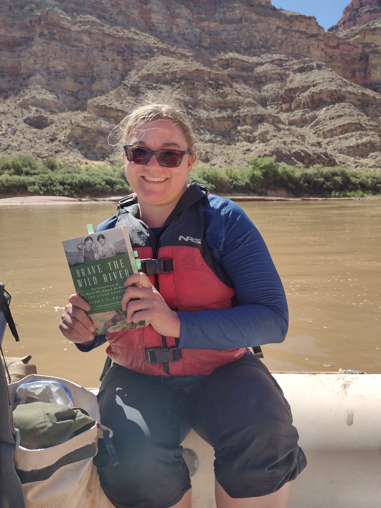 A white woman with blonde hair and sunglasses wearing a life jacket, perched on the edge of a boat, holding up a book. There are steep, rugged desert cliffs beyond the river. It' a sunny day.