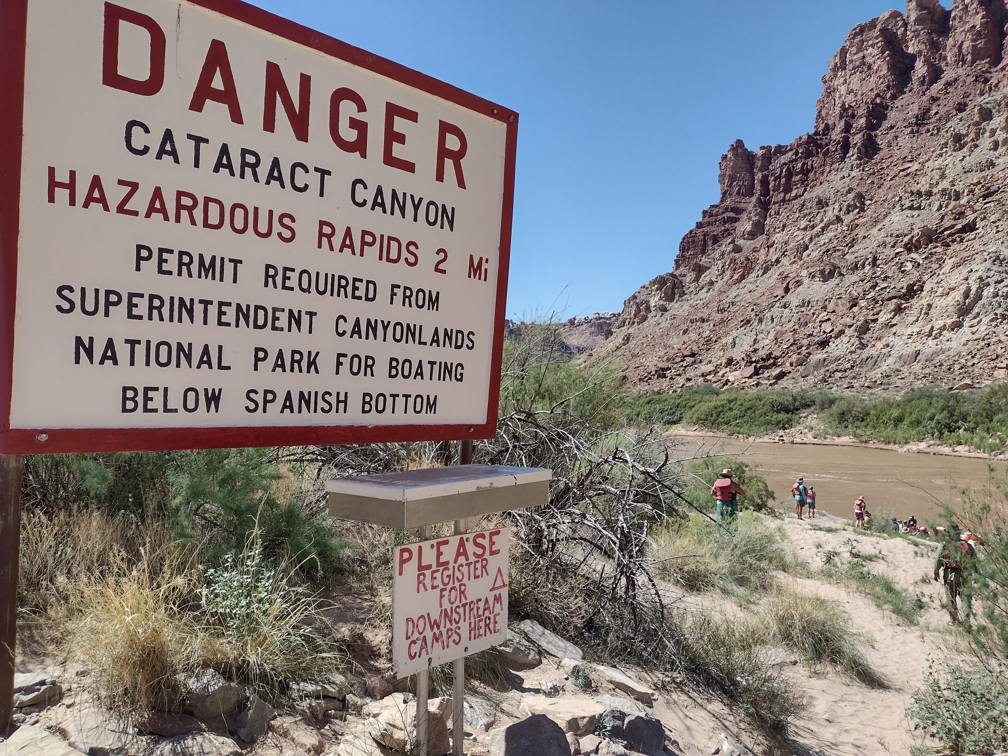 A photo of a sign with red lettering saying DANGER: Cataract Canyon Hazardous Rapids 2 mi. Below that is a metal box and a sign asking people to register for a campsite. In the distance, a river below steep, rugged cliffs, and a few people walking toward it.