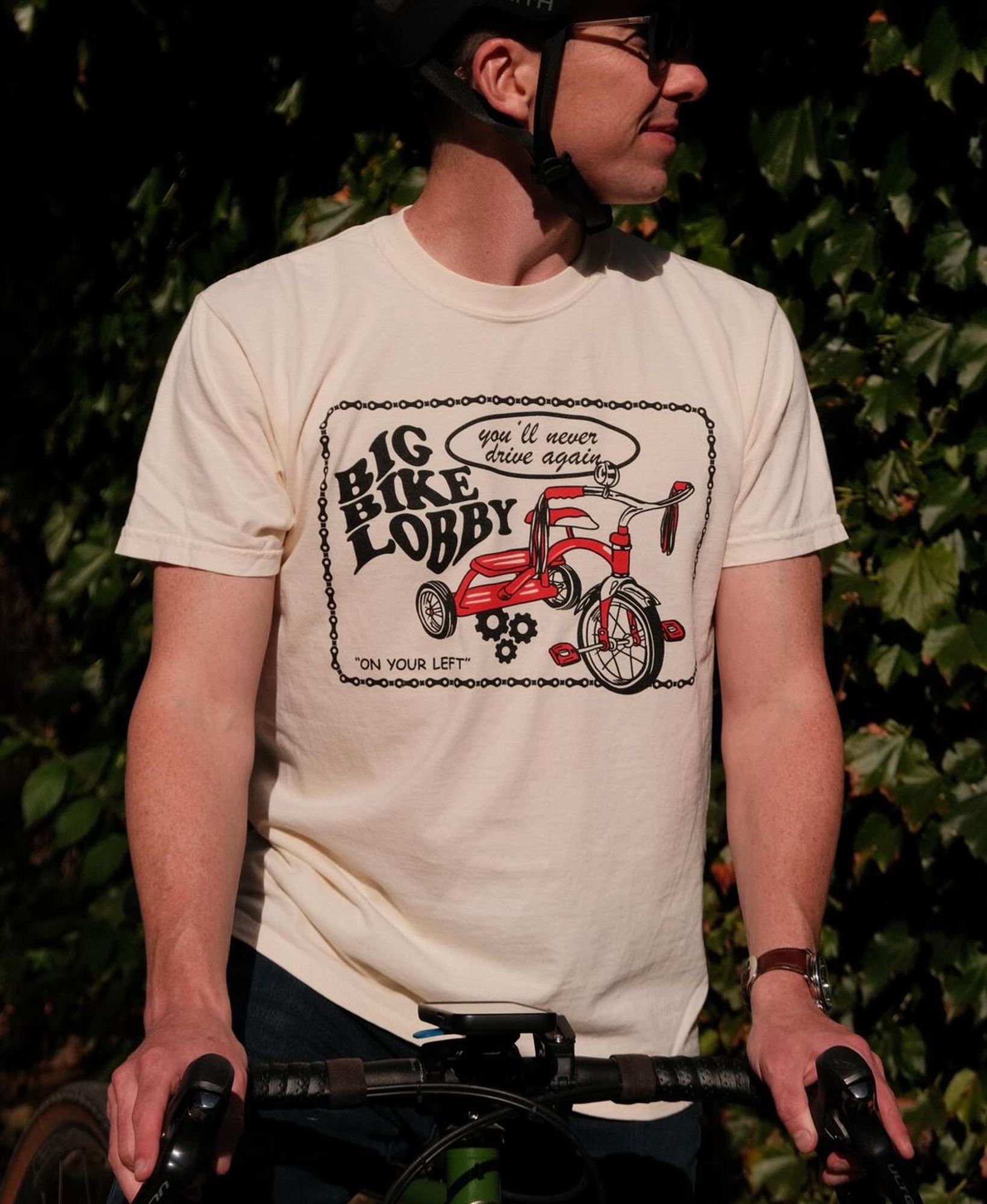 Person facing front with a shirt that says "Big Bike Lobby" with a classic red tricycle. There's "on your left!" and "you'll never drive again" in smaller font surrounded by a bike chain border