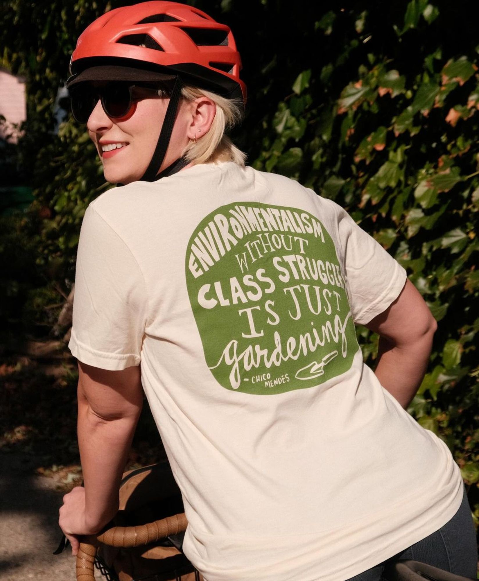 Person wearing a shirt that says "Environmentalism without class struggle is just gardening - Chico Mendes" with a drawing of a spade. the design is on the back, the person is wearing a helmet on a bicycle