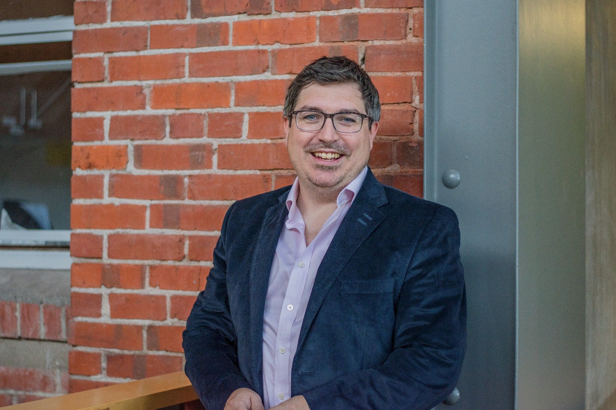 SOJC journalism Professor of Practice Damian Radcliffe poses for a photo in front of a brick wall.